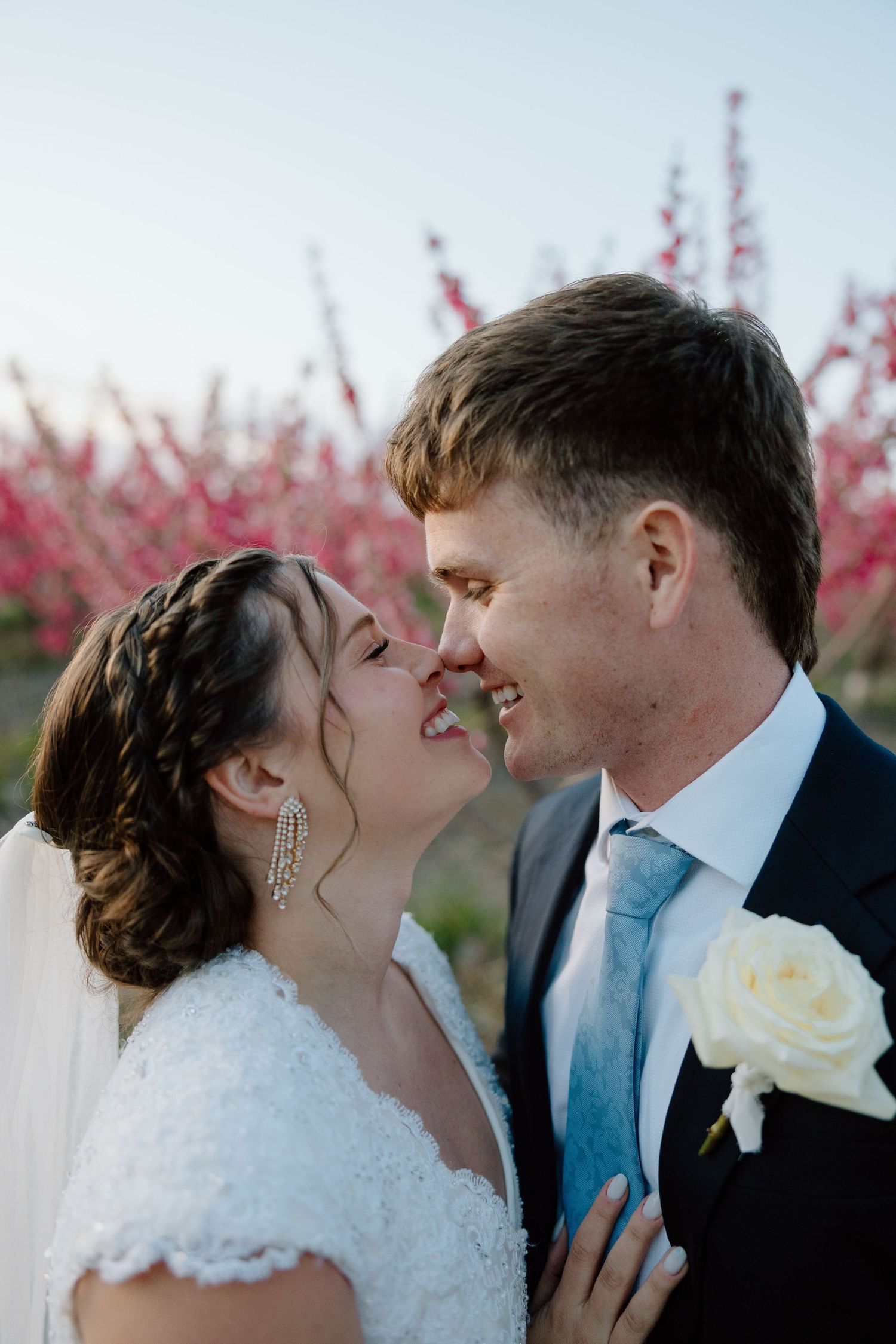 Bride and groom share a joyful nose-to-nose moment in front of vibrant pink blossoming trees.