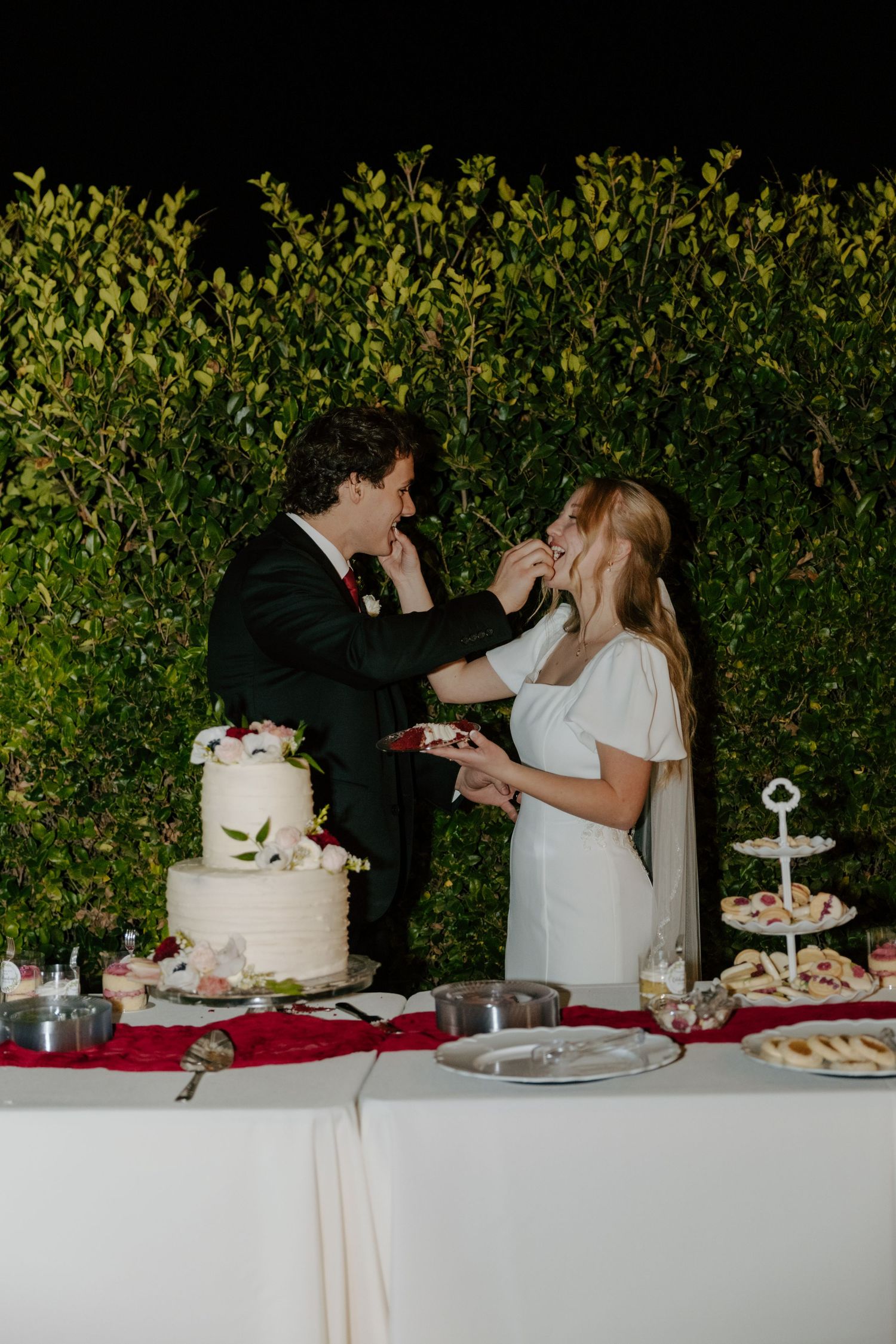 Bride and groom feed each other wedding cake at dessert table in front of green hedge backdrop.