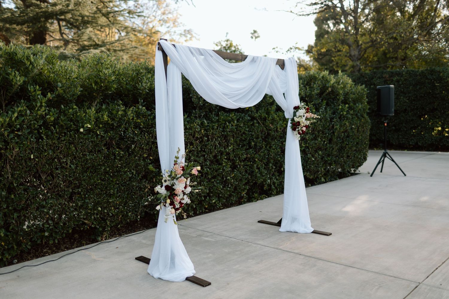 Elegant outdoor wedding arch draped in white fabric with pink floral arrangements on a patio surrounded by green hedges.