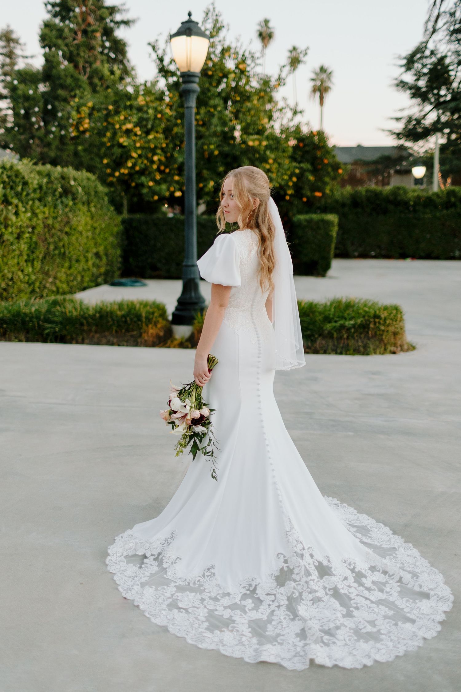 Bride in white flutter-sleeve mermaid wedding gown with lace train holds bouquet turning back to camera outdoors.