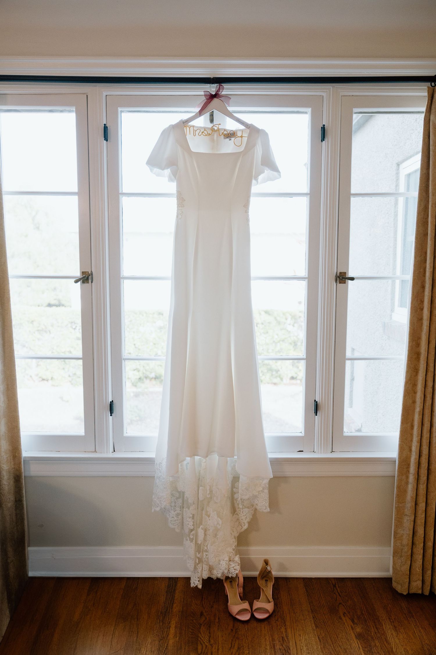 White short-sleeve wedding gown with lace hem hangs in a sunlit window above nude strappy bridal heels.