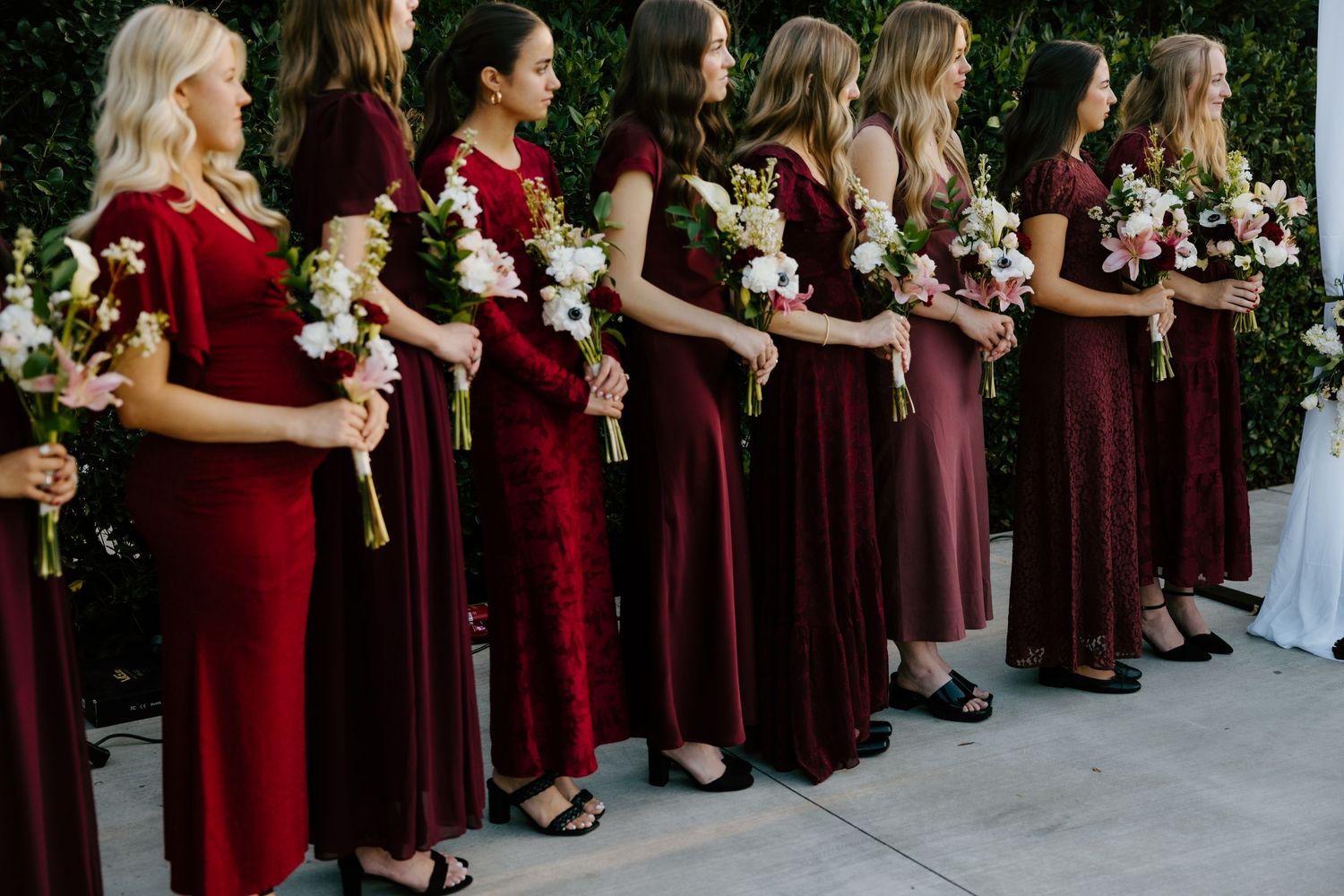 Row of bridesmaids in burgundy velvet dresses holding white and blush bouquets at an outdoor wedding.