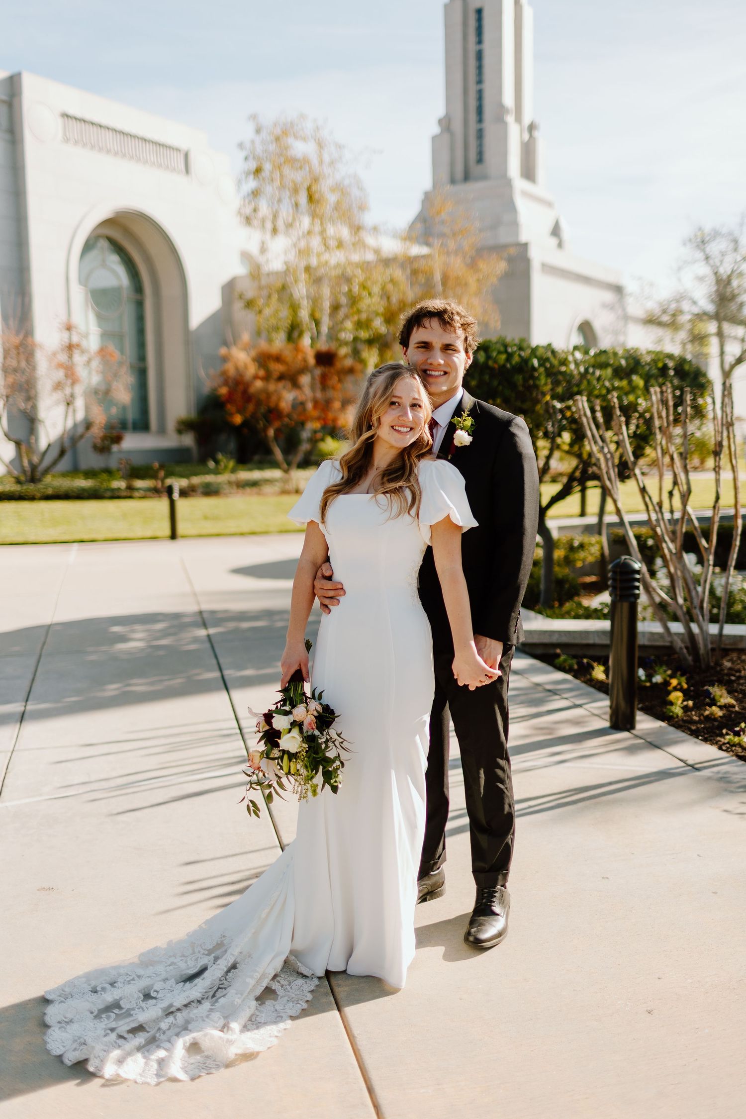 Bride and groom posing together outside a white temple on their wedding day, bride holding a floral bouquet.