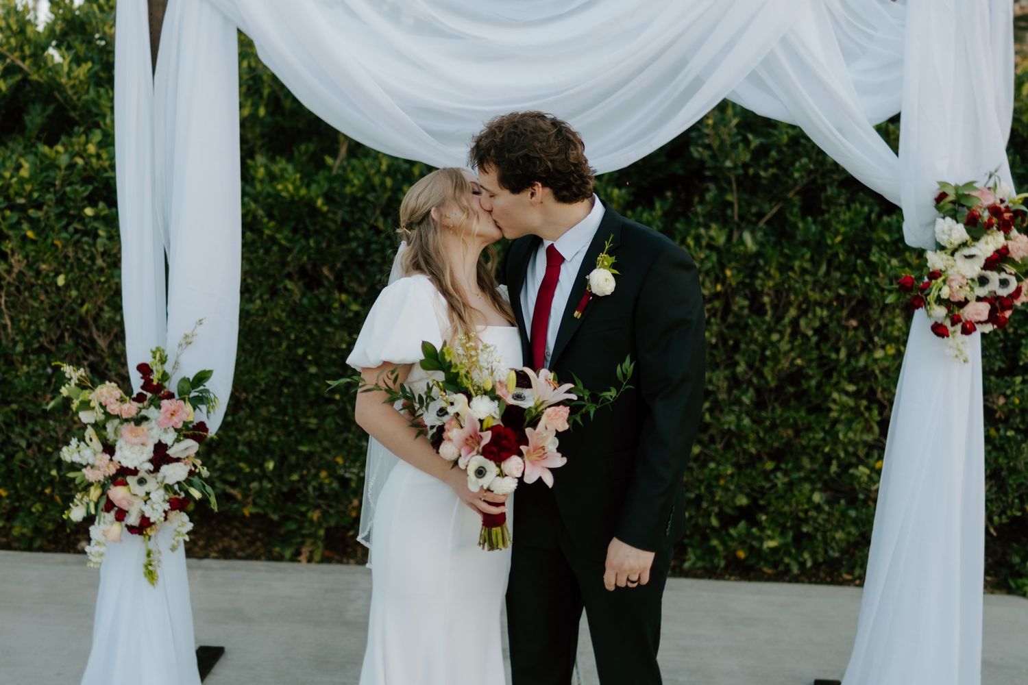 Bride and groom sharing a kiss under a white draped wedding arch adorned with pink and red floral arrangements.