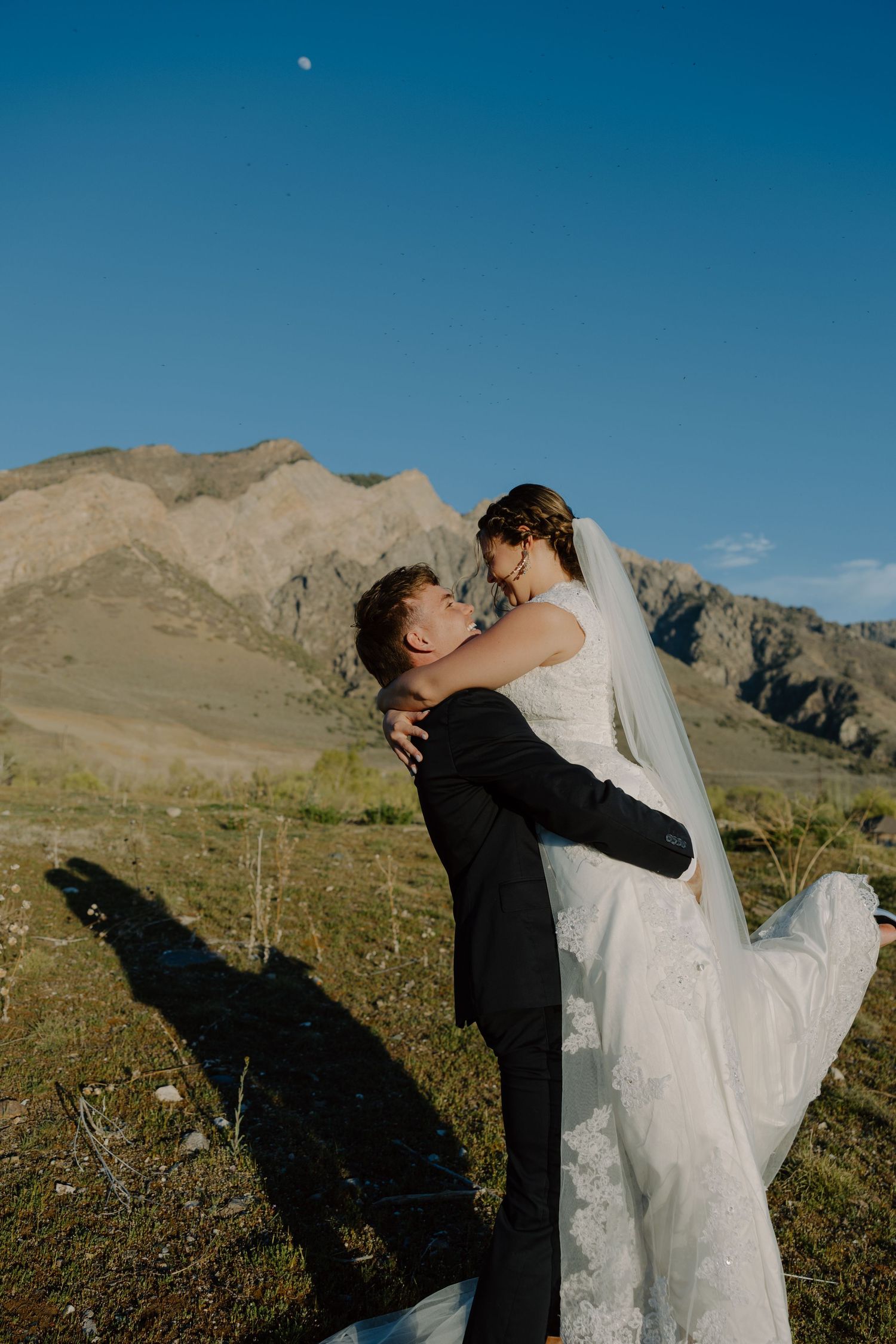 Groom lifting smiling bride in white wedding dress with veil flowing in mountain setting under blue sky.