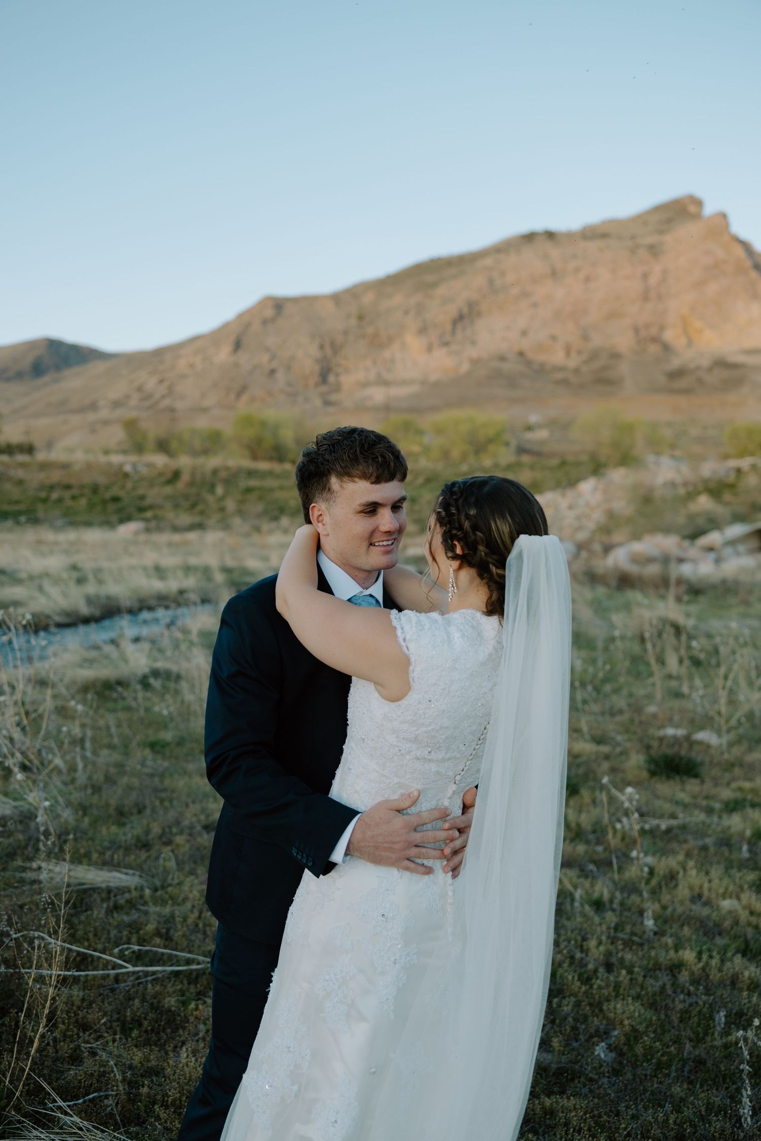 Smiling groom holds his bride close in an outdoor wedding portrait with a dramatic mountain backdrop.