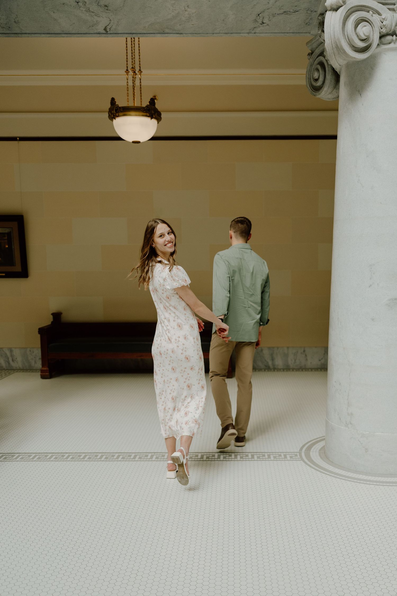A woman in a floral dress smiles back while holding hands with a man walking through an elegant marble hall.