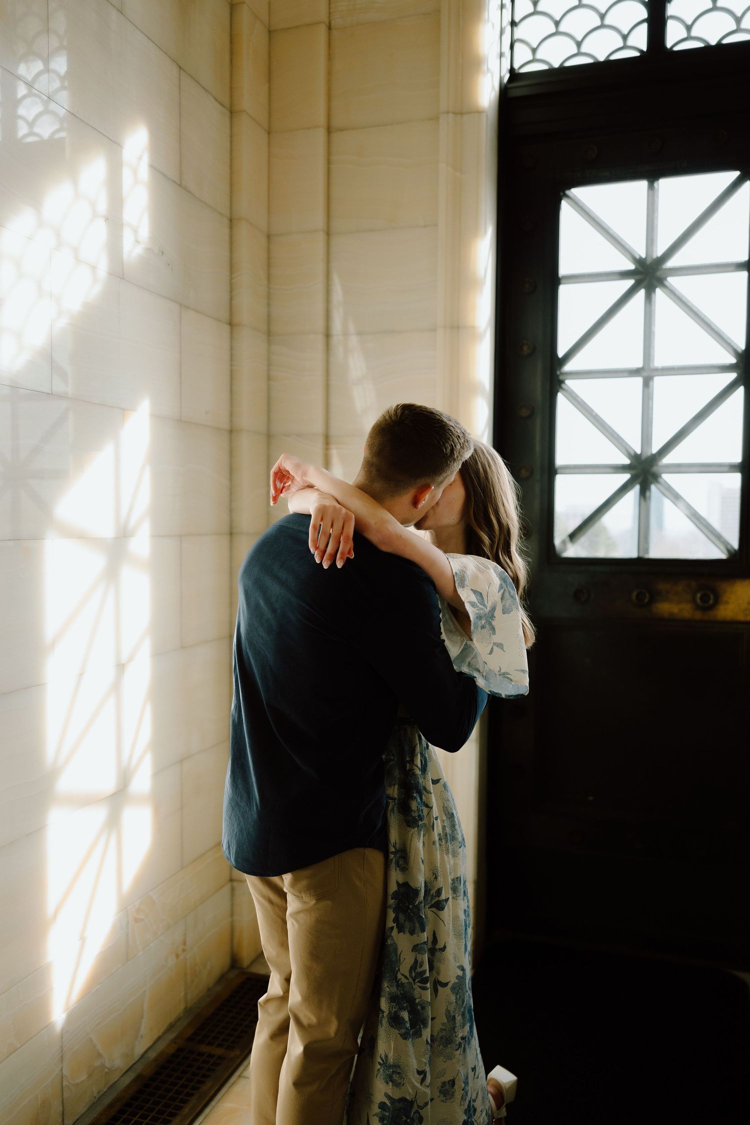 Couple sharing a tender kiss near an ornate dark door with sunlight streaming through a latticed window.