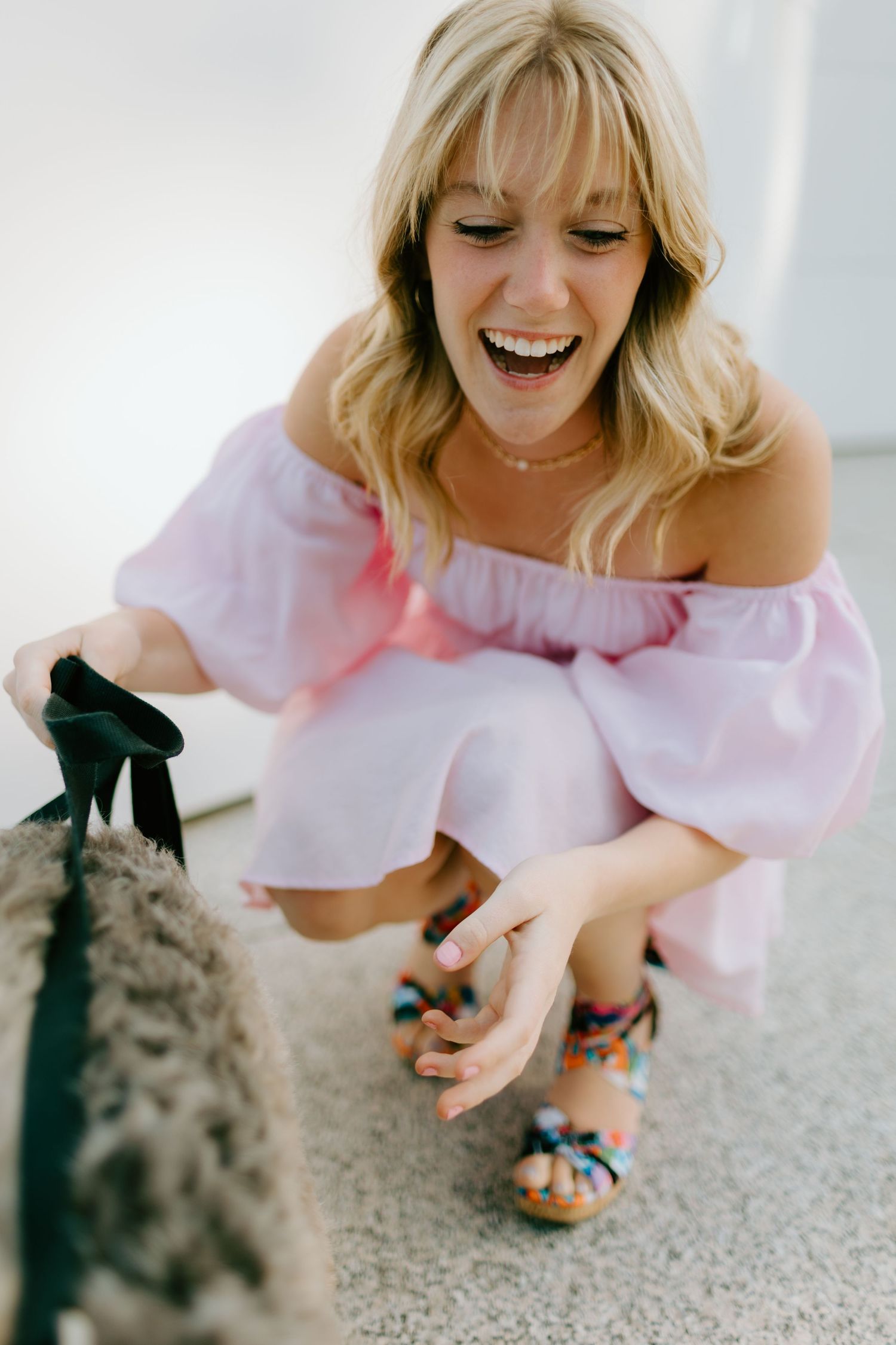 Smiling blonde woman in pink off-shoulder dress and colorful sandals crouching down to pet a fluffy dog.
