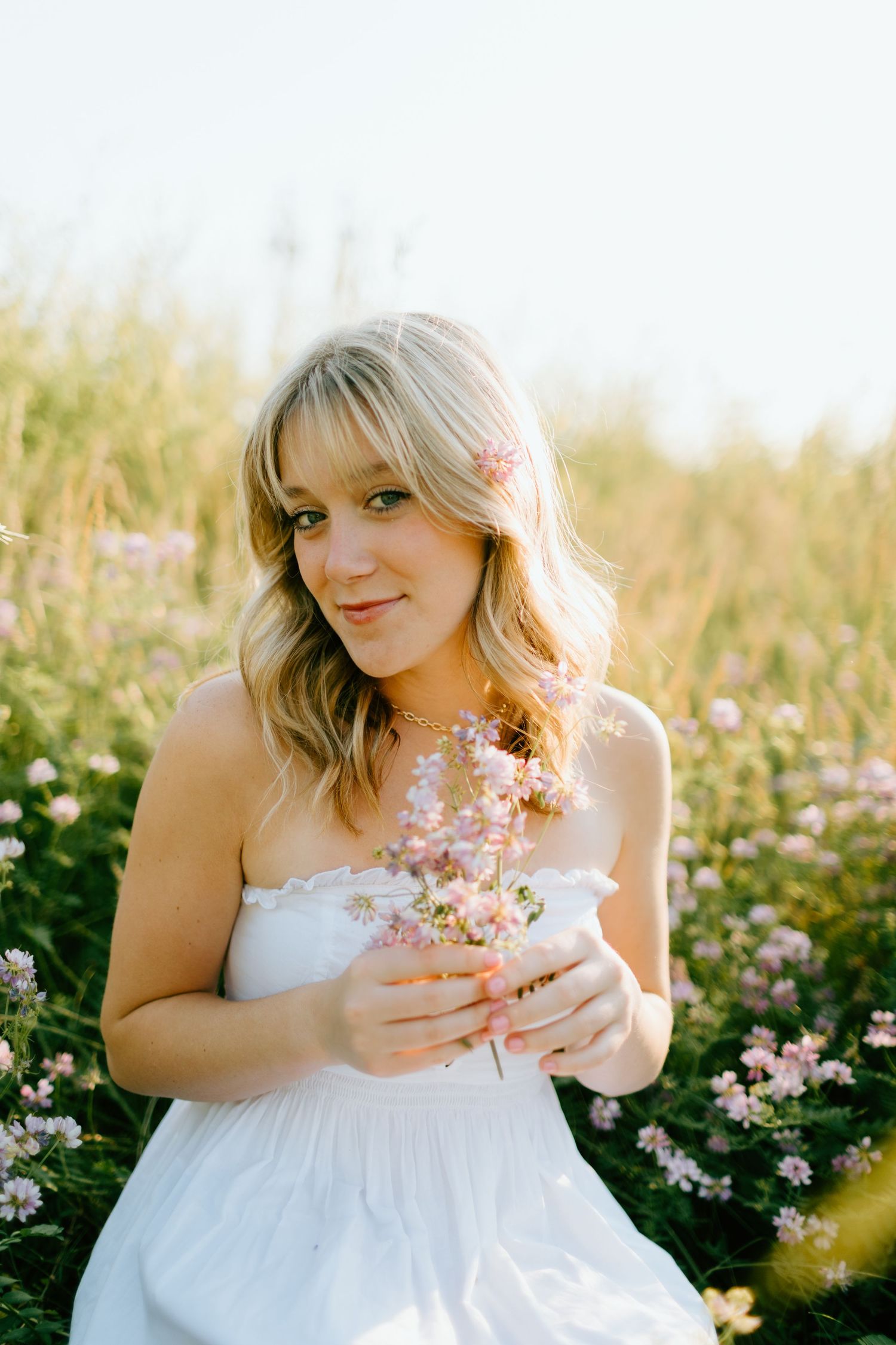 Young blonde woman in white dress holding pink wildflowers in a sunlit meadow field.