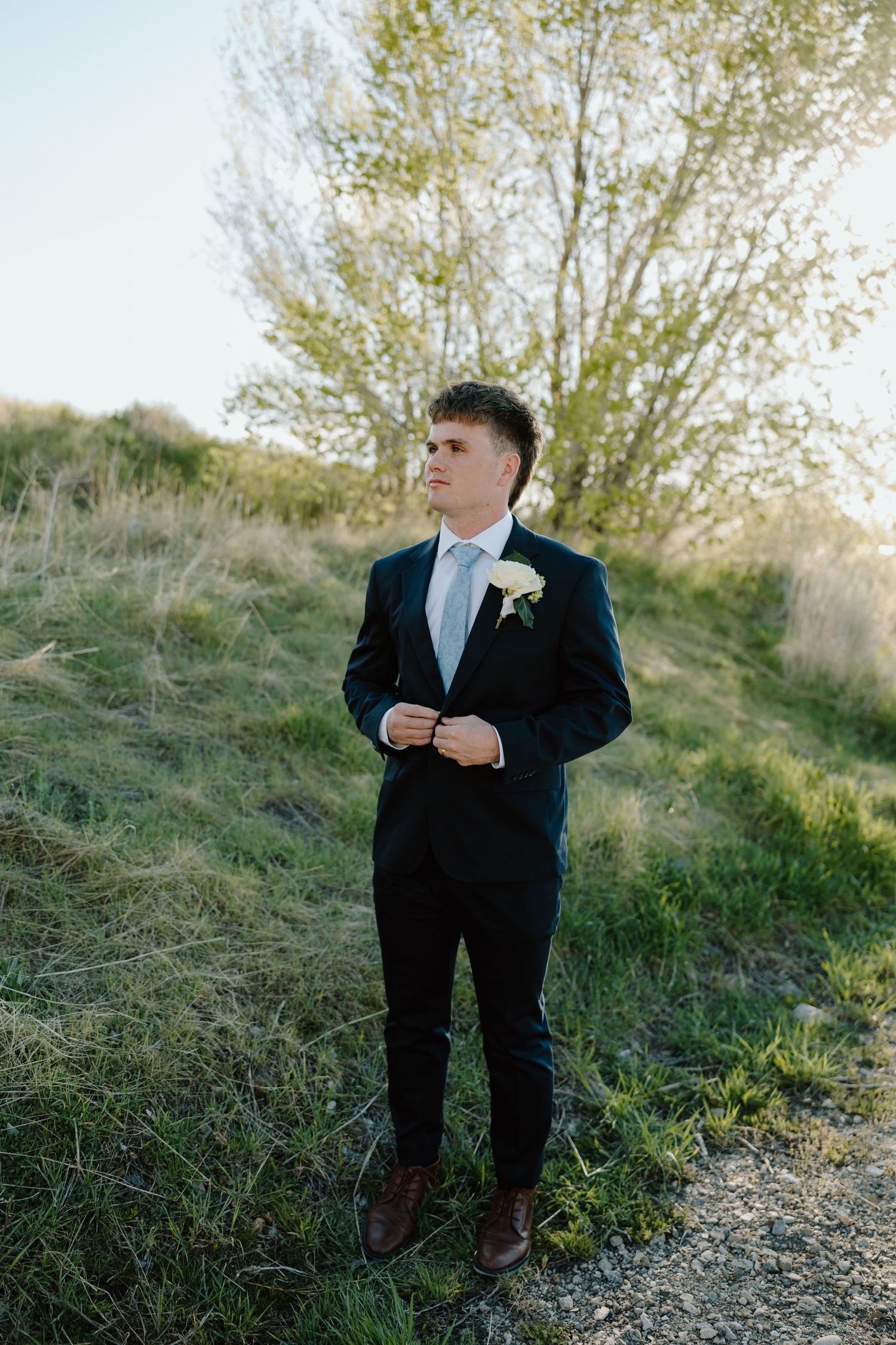 Groom in dark suit with white boutonniere adjusting jacket outdoors in green field.