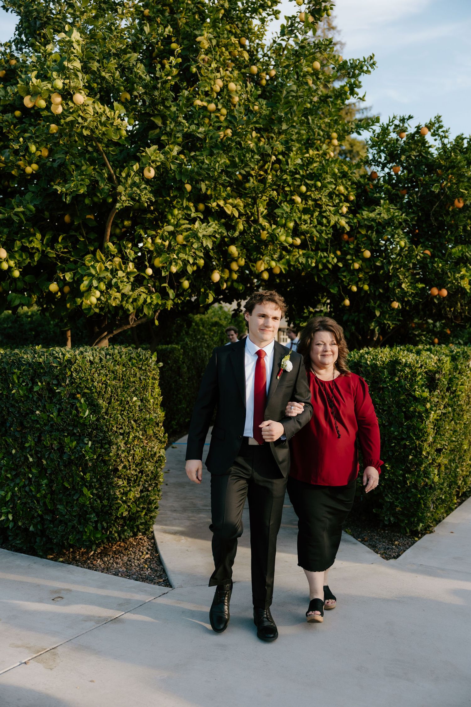 Groom in dark suit and red tie walks with mother through garden hedge pathway at outdoor wedding.