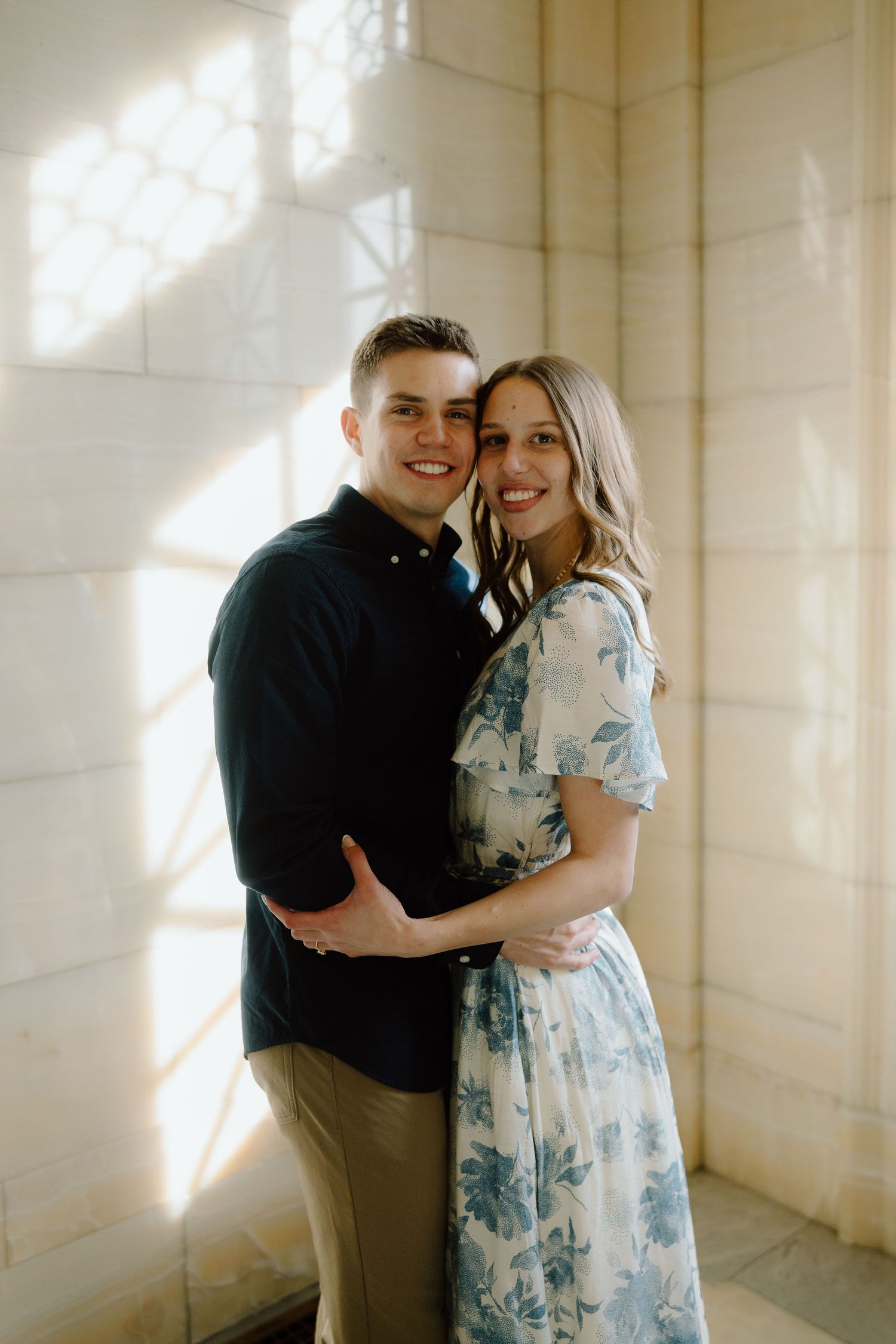 Happy couple embracing and smiling in warm sunlight against a stone wall, woman in floral dress.