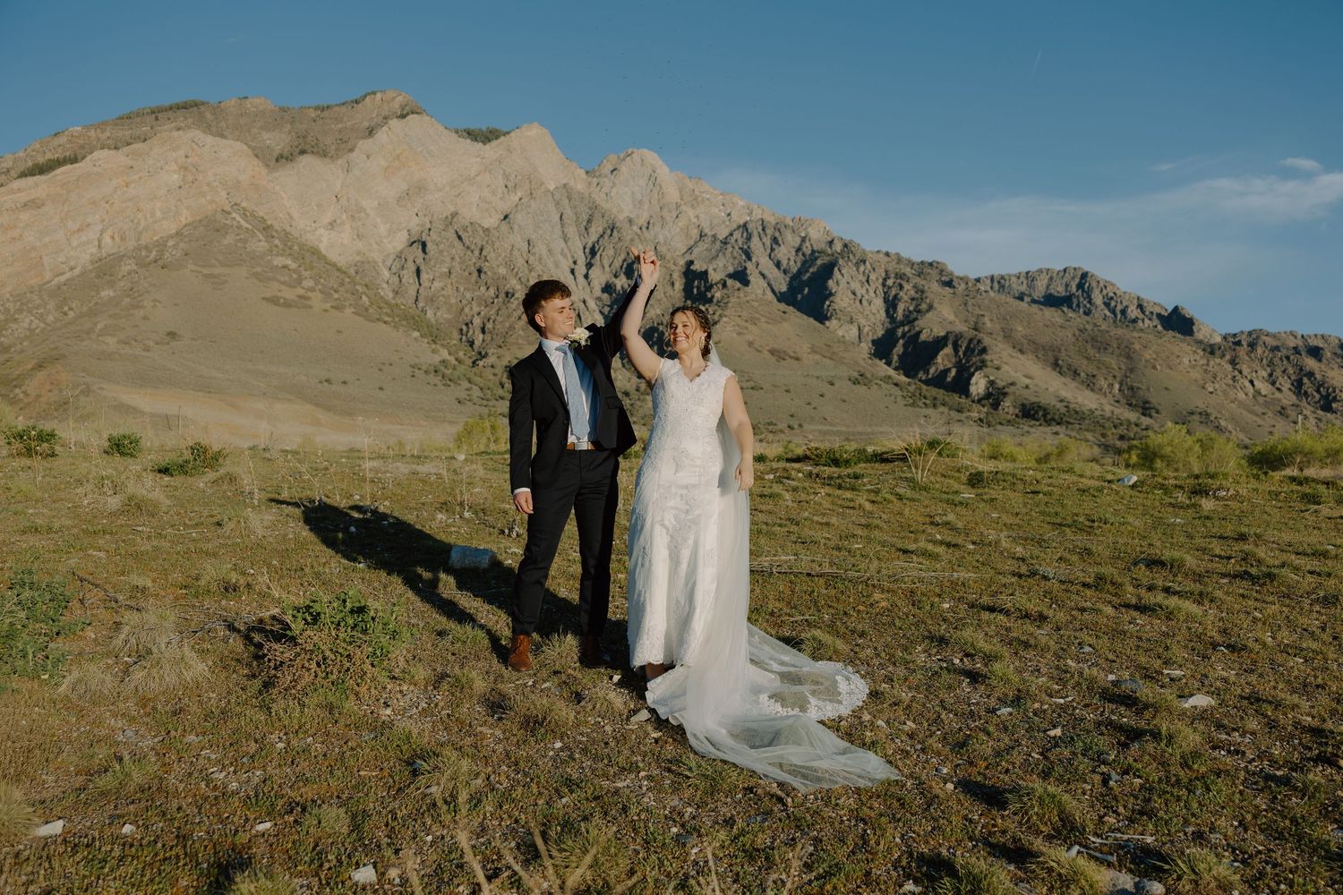 Joyful bride and groom raise hands in celebration with dramatic mountain landscape behind them.