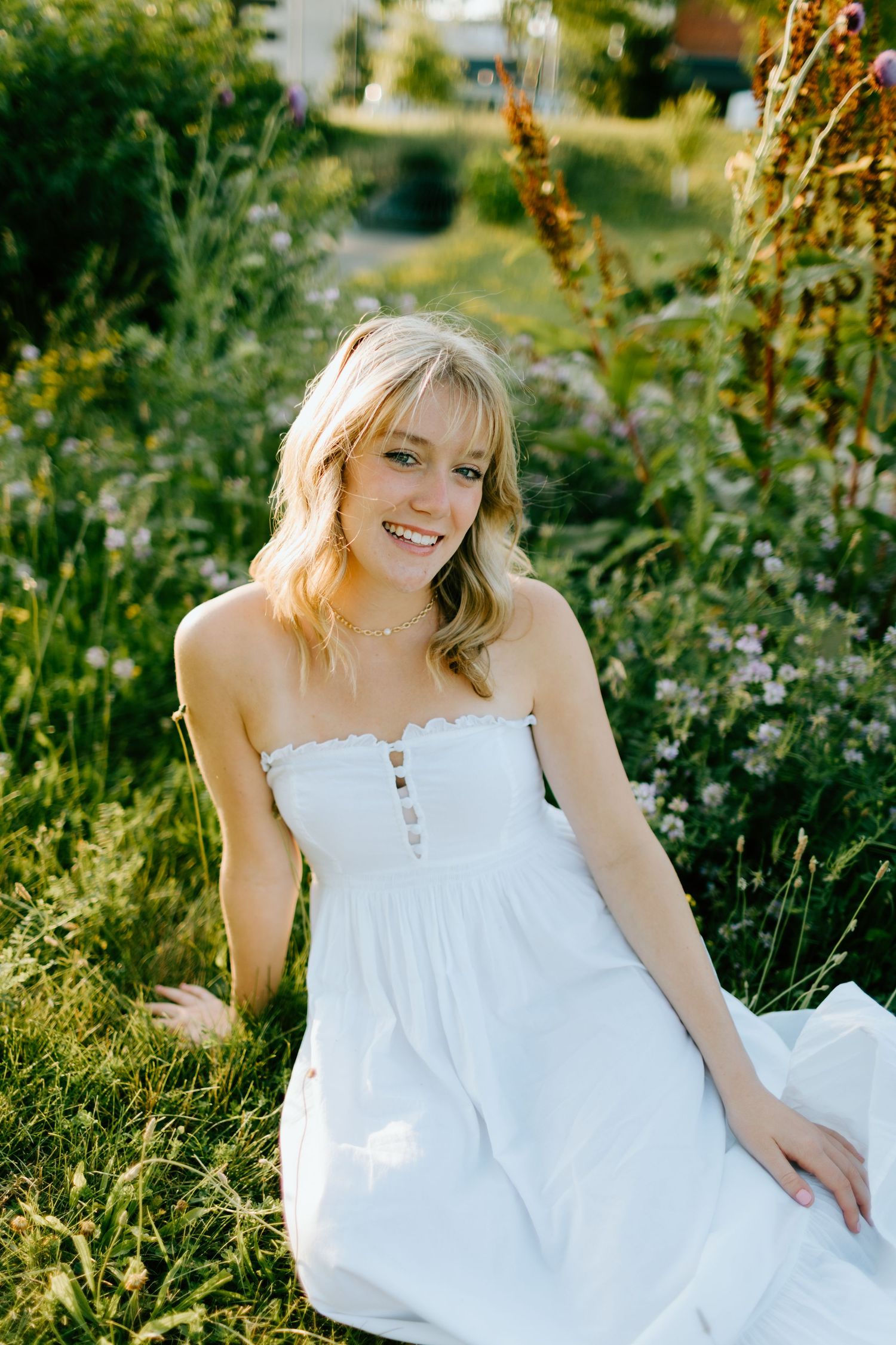Smiling blonde woman in white strapless sundress sitting in lush green garden surrounded by wildflowers in golden light.