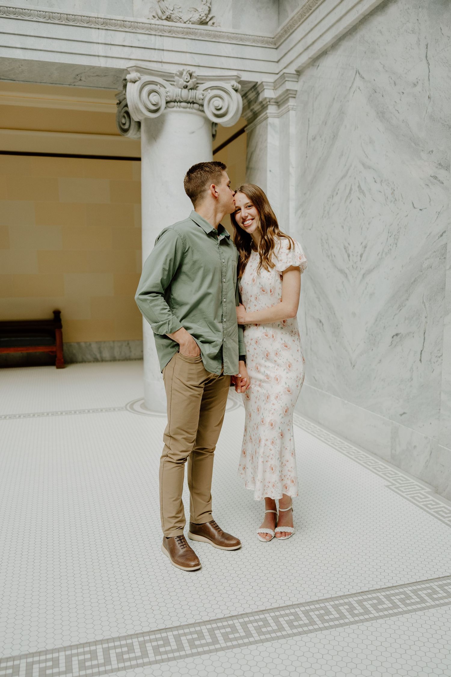 Couple sharing a romantic moment in an elegant marble building, man in green shirt kissing woman in floral dress.