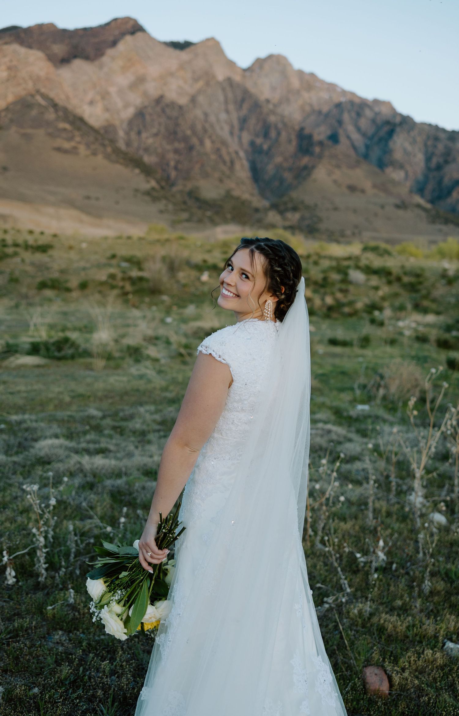 Smiling bride in white lace gown holds bouquet, posing before scenic desert mountains at golden hour.