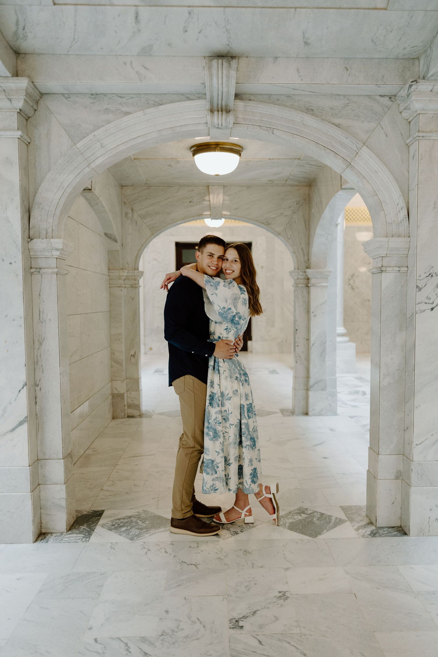 A smiling couple embraces tenderly beneath a marble archway in a grand classical building interior.