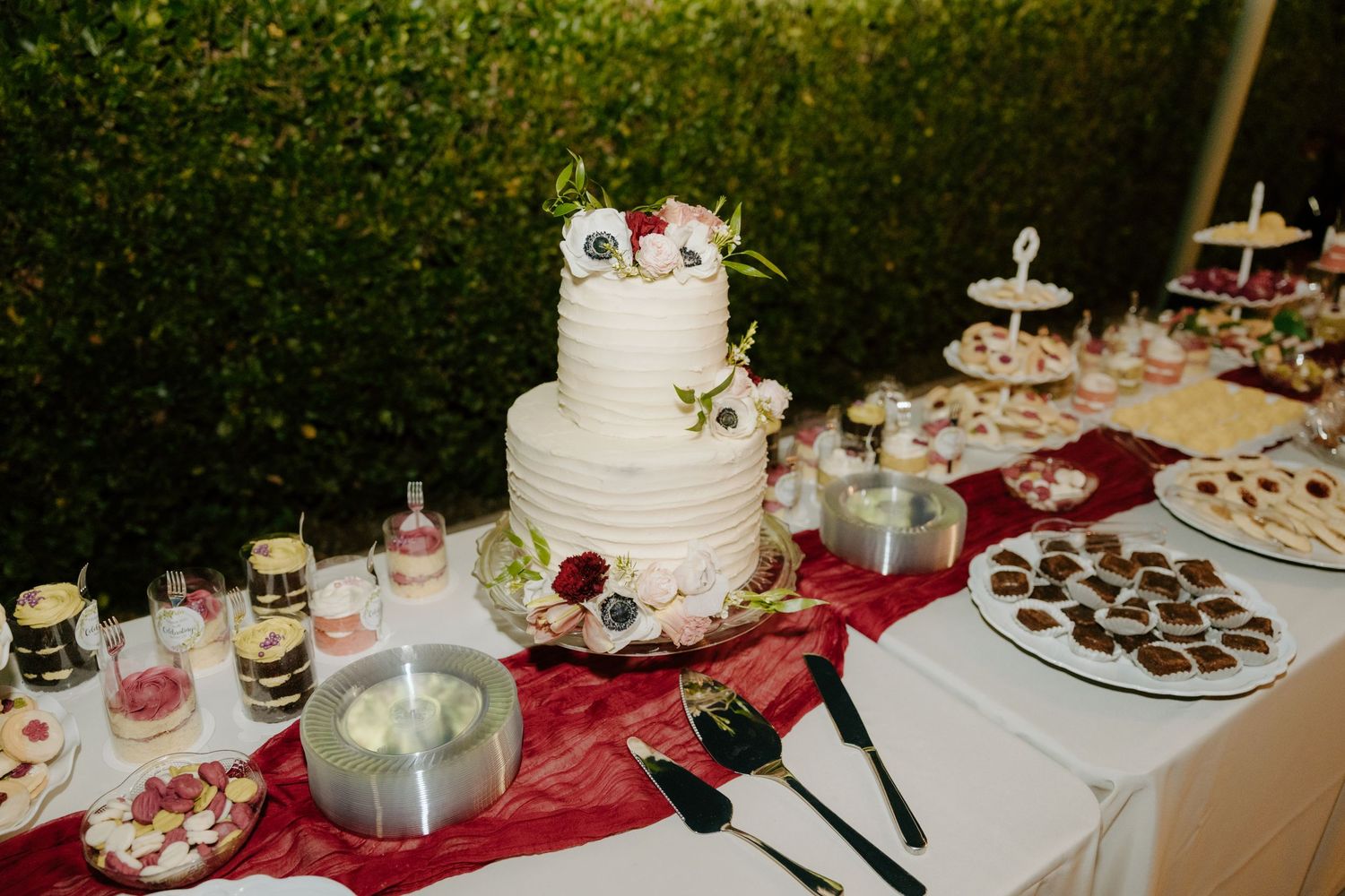 Elegant white three-tier wedding cake surrounded by desserts on a burgundy table runner.