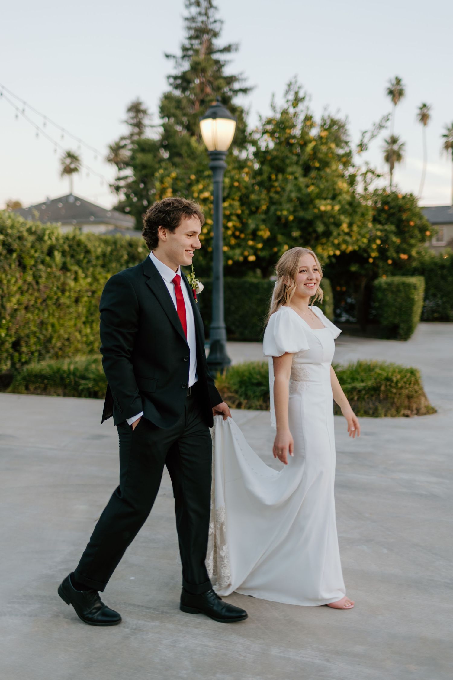 A bride in a white flutter-sleeve gown and groom in a black suit with red tie walk together outdoors near palm trees.