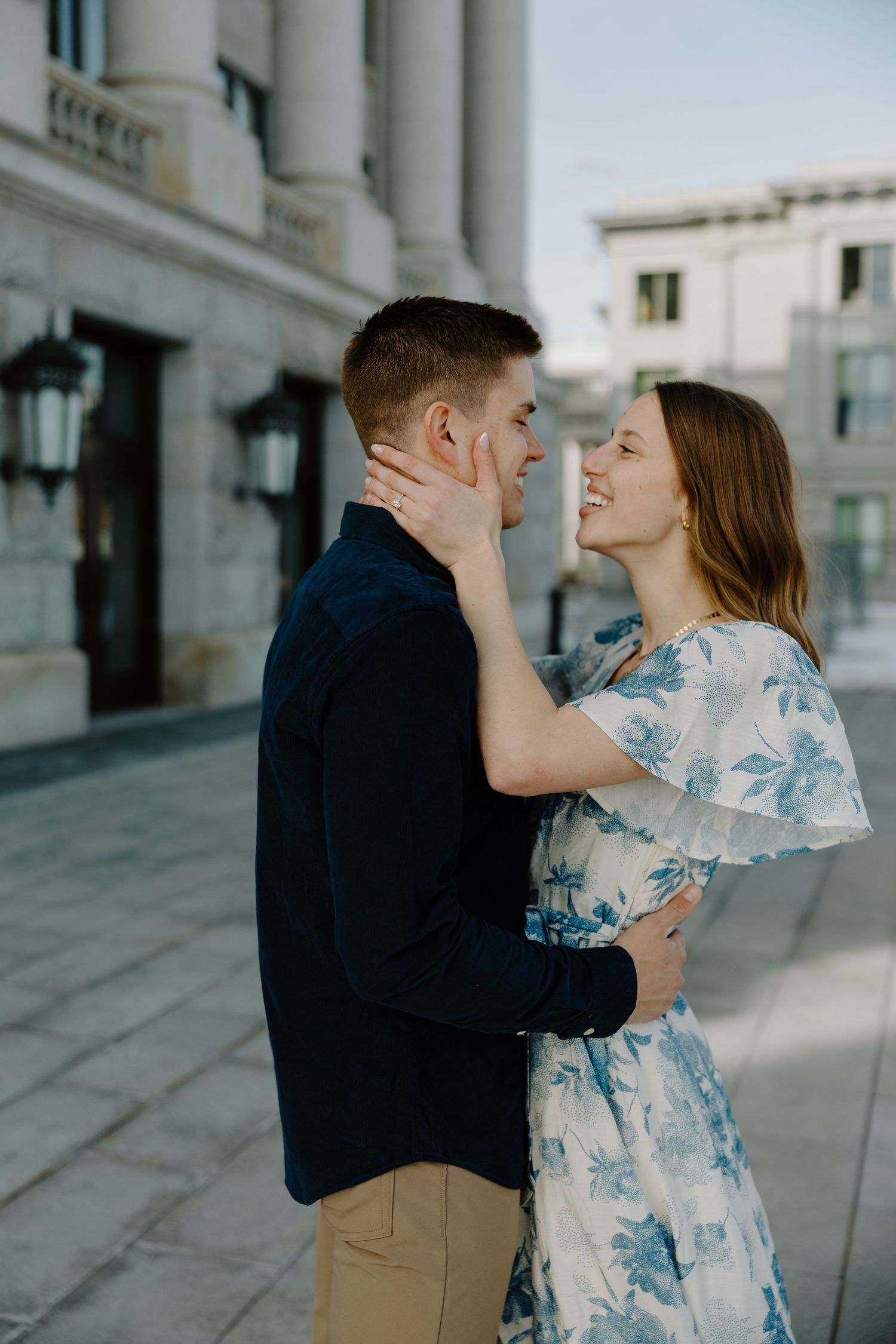 Smiling couple embracing outdoors, woman in blue floral dress tenderly holding mans face near classic building.