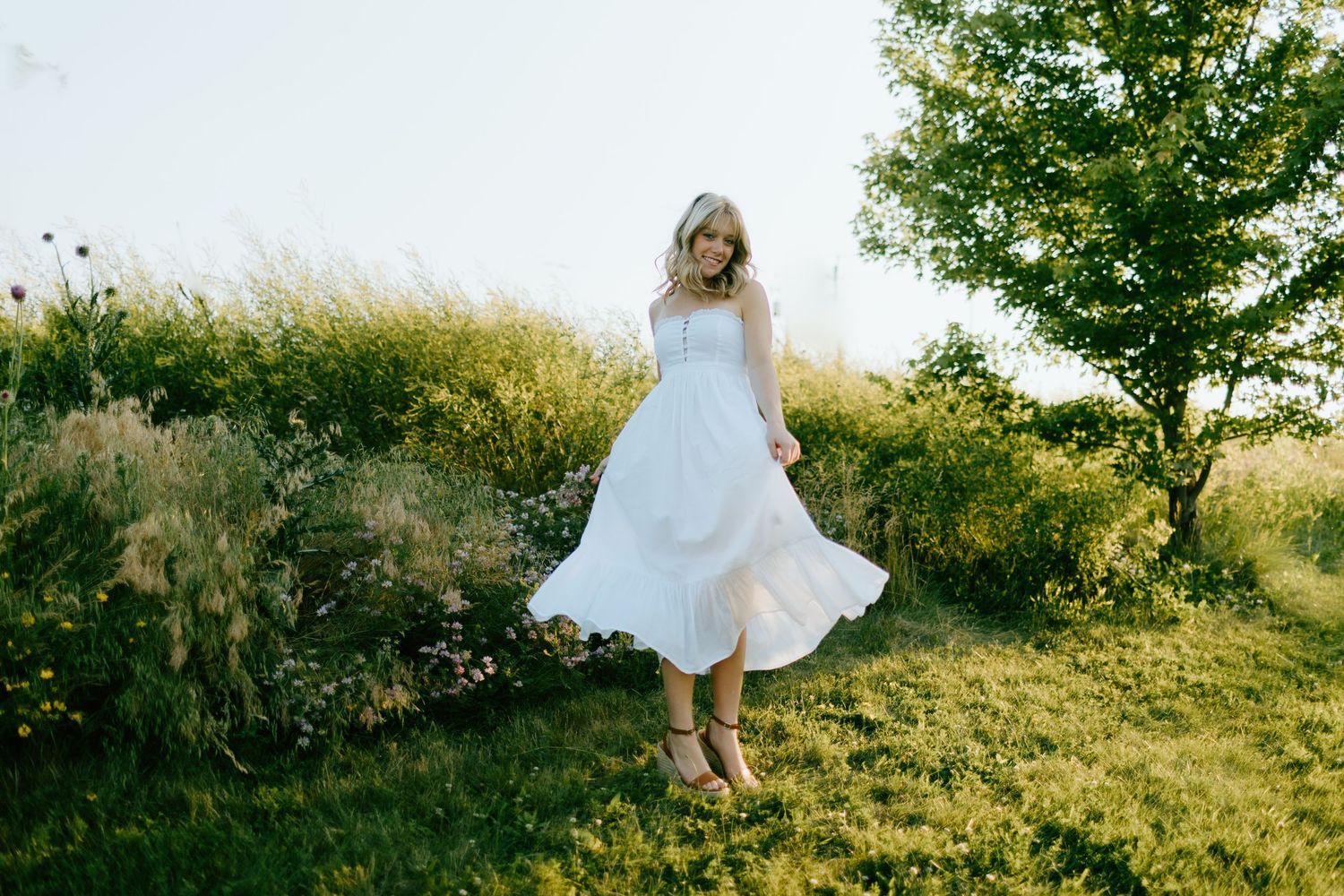 Woman in white midi dress twirling in lush green garden with wildflowers and trees on sunny day.
