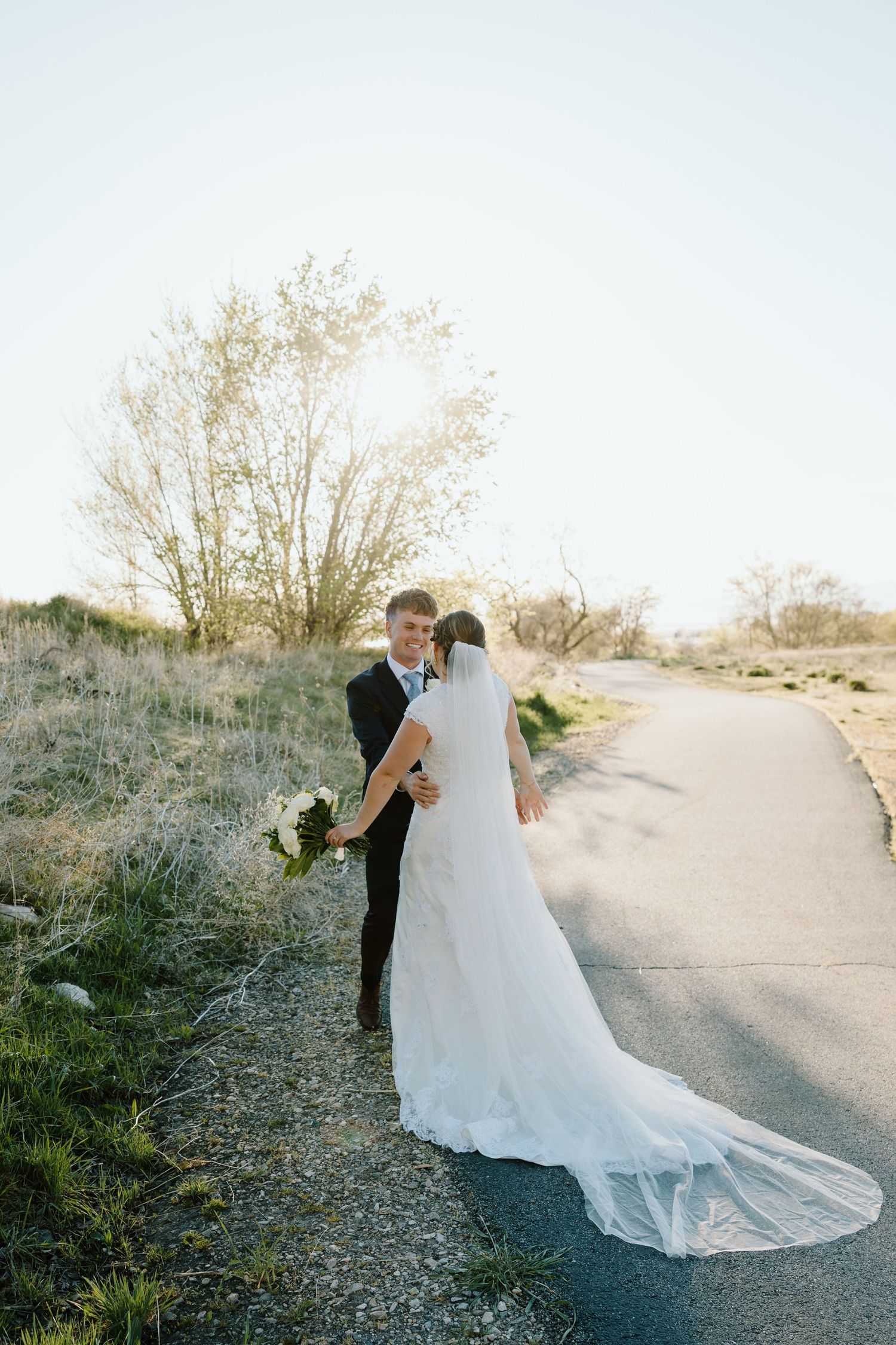Bride and groom embracing on a sunlit path surrounded by nature in a romantic wedding portrait.