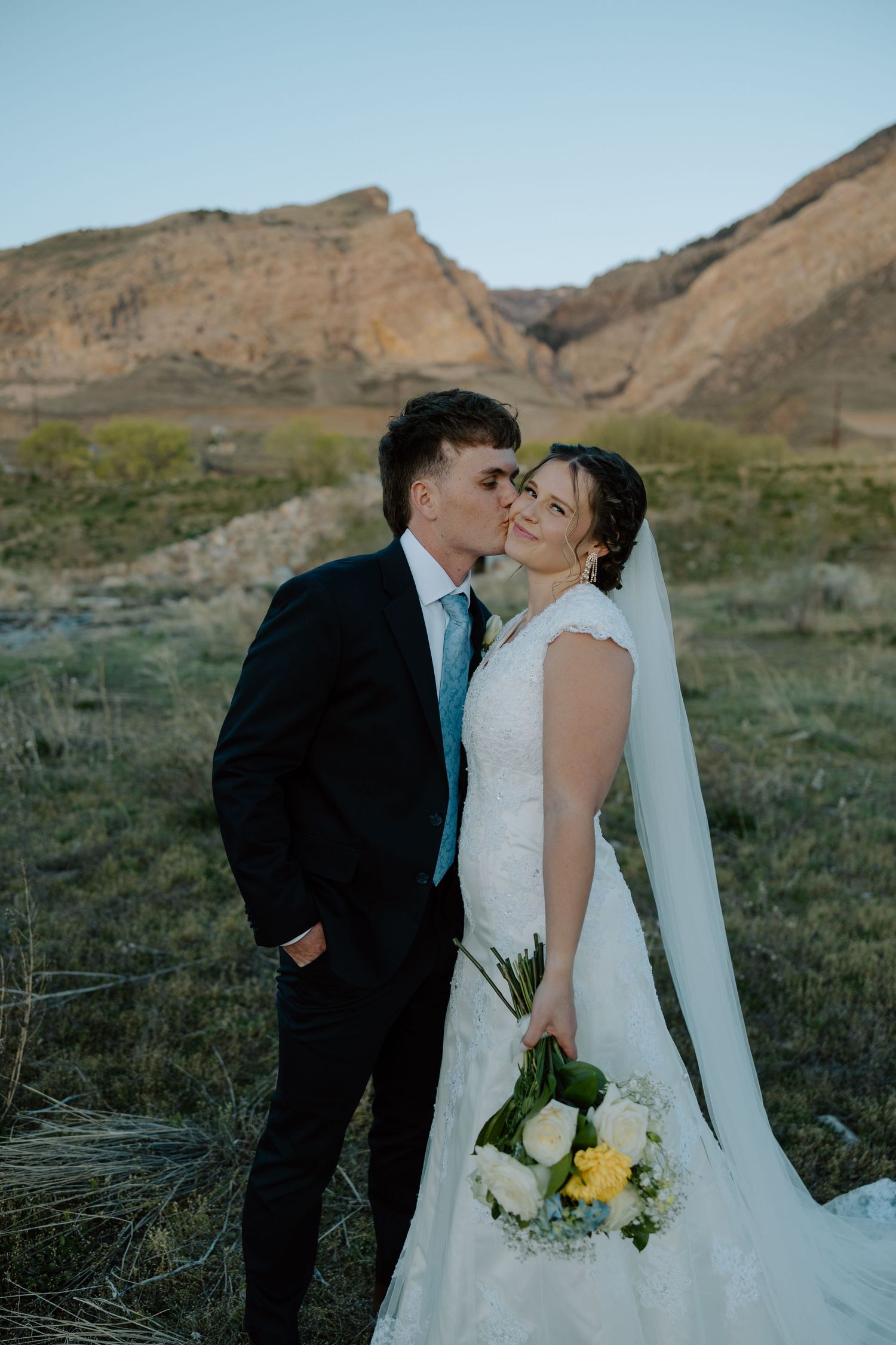 Groom kisses bride on the cheek as she holds a white and yellow bouquet with mountains behind them.