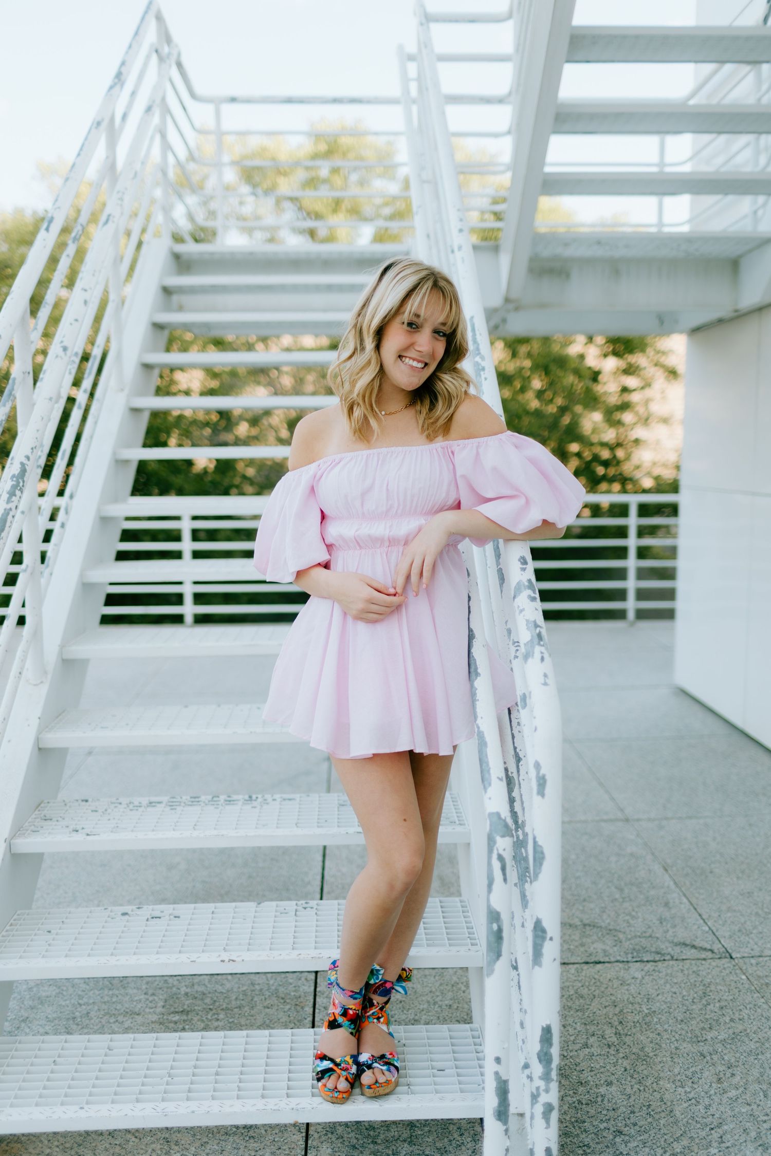 Woman in pink off-shoulder mini dress and colorful heeled sandals smiling on white outdoor stairs.