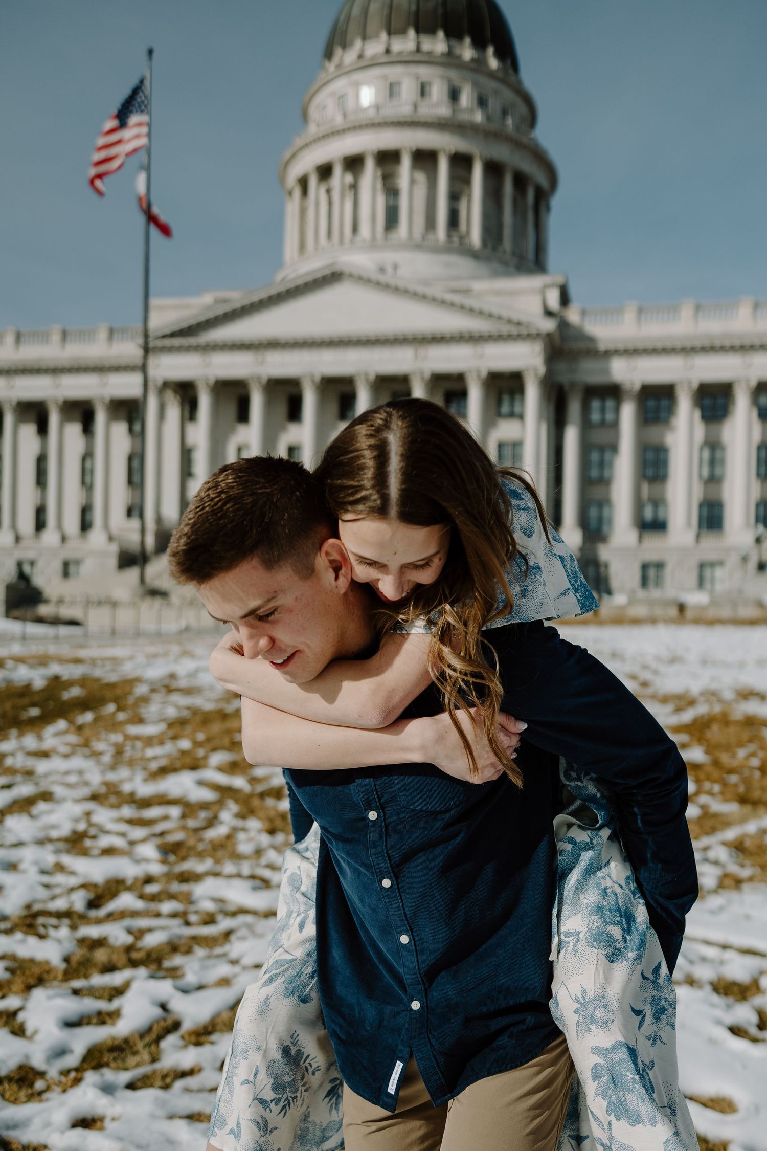 Couple sharing a piggyback ride in front of the Utah State Capitol building with snow on the ground.