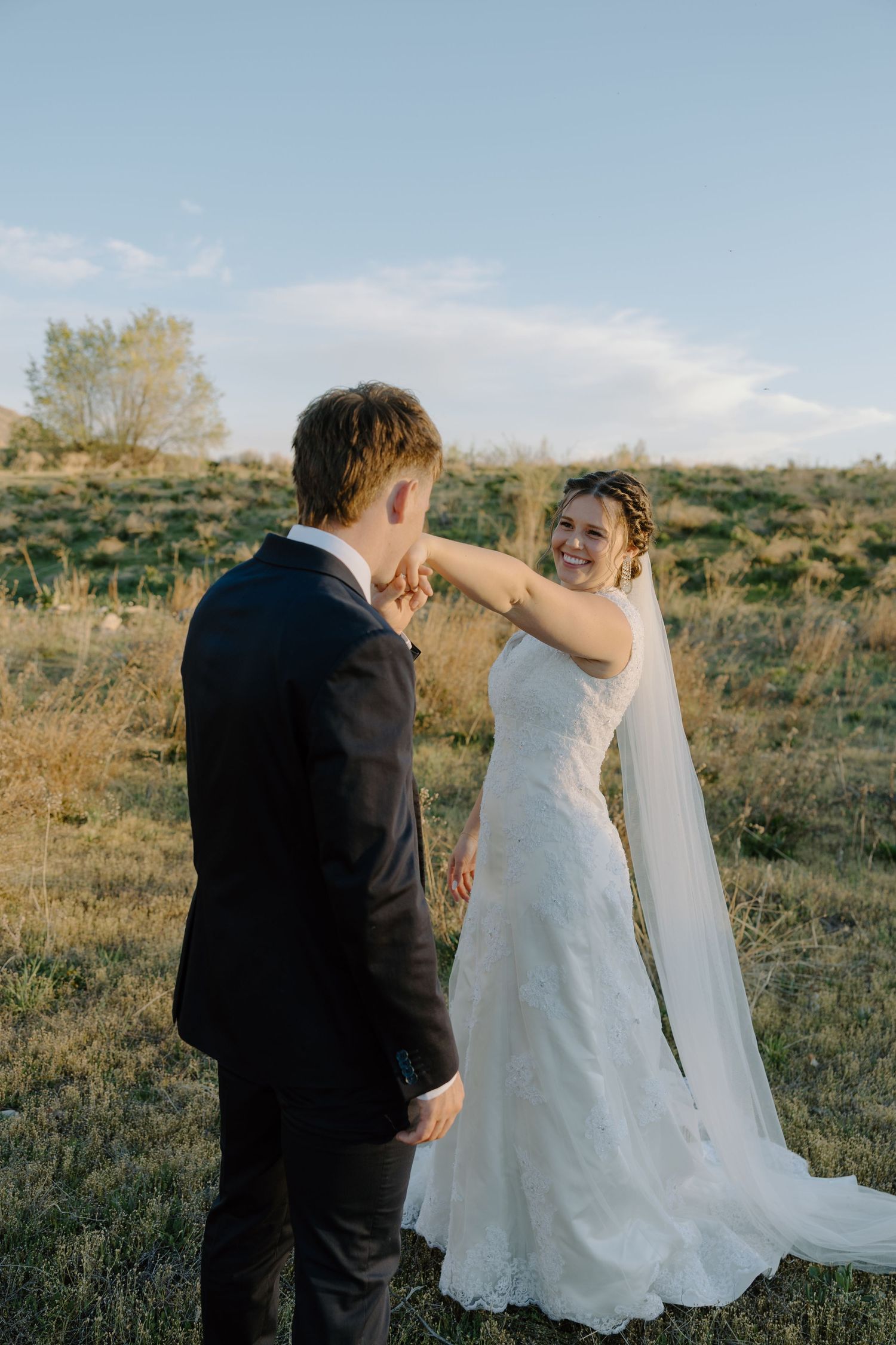Bride in white lace gown and veil laughs with groom in black suit during outdoor wedding portraits at golden hour.