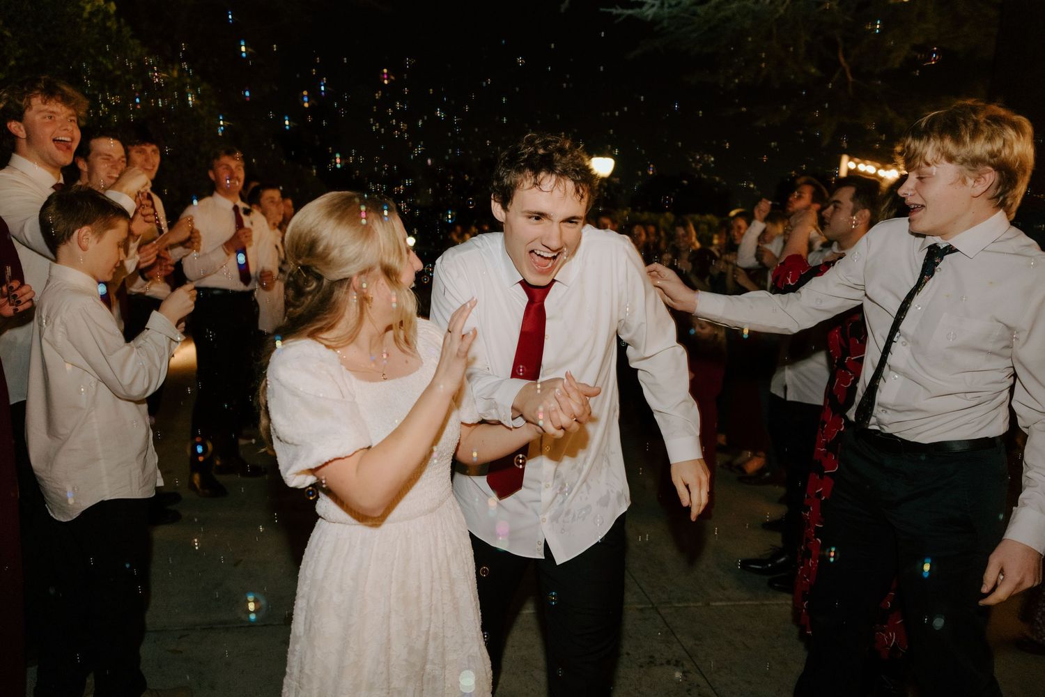 Joyful couple dancing at wedding reception surrounded by cheering guests at night outdoor celebration.