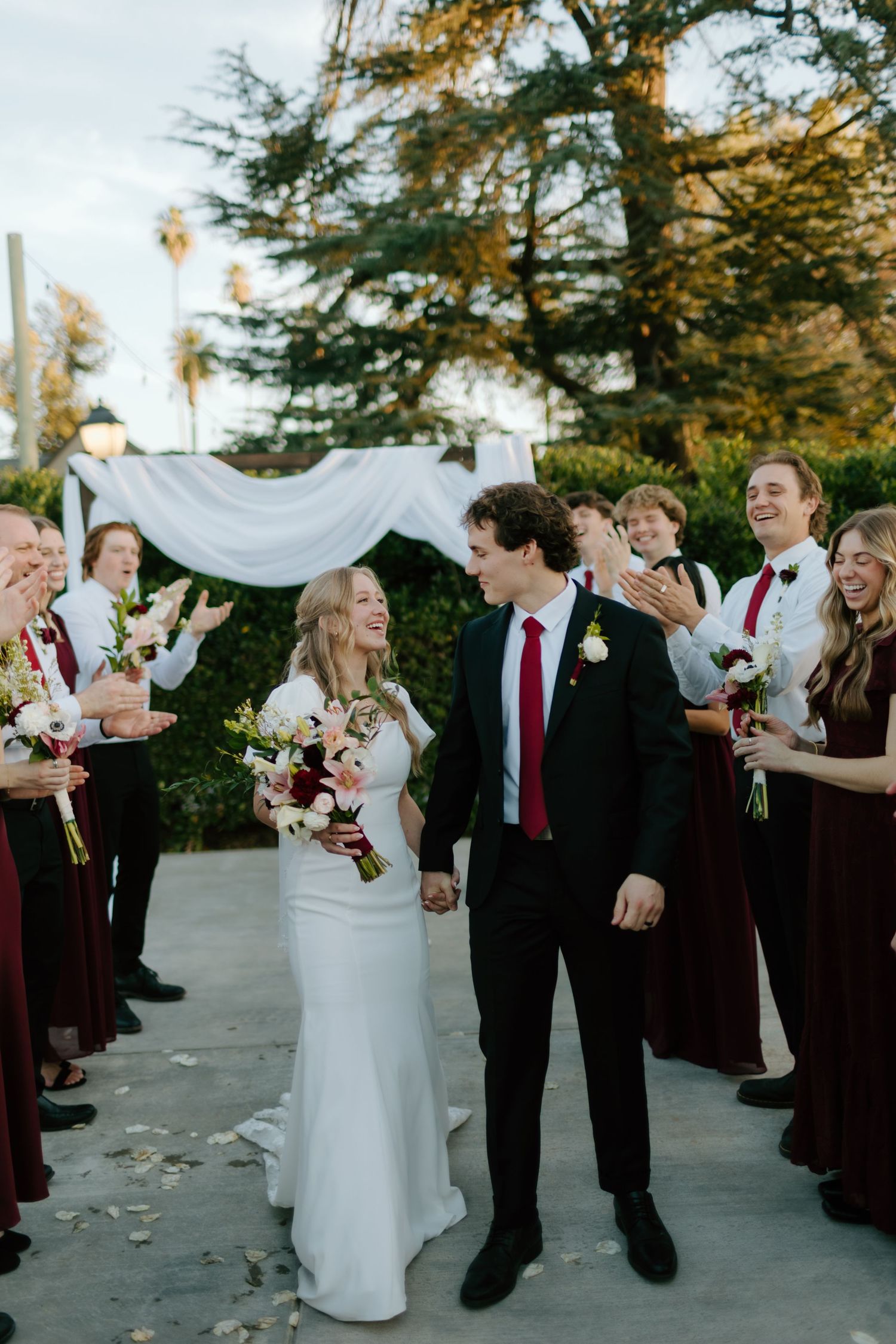Newlyweds walk through cheering wedding party in burgundy attire after outdoor ceremony.