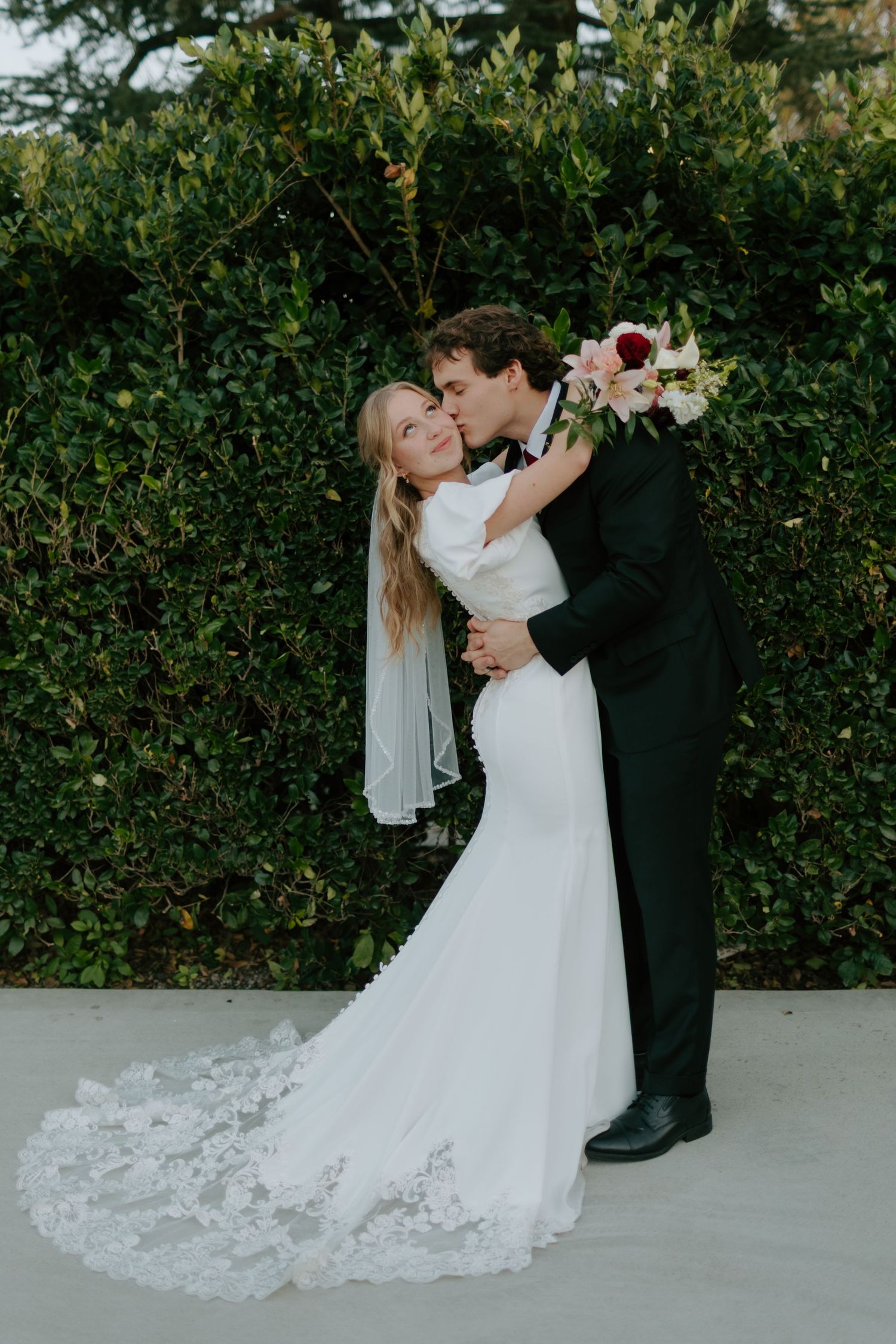 Groom in black tuxedo kisses bride in lace wedding gown as she smiles before a lush green hedge.