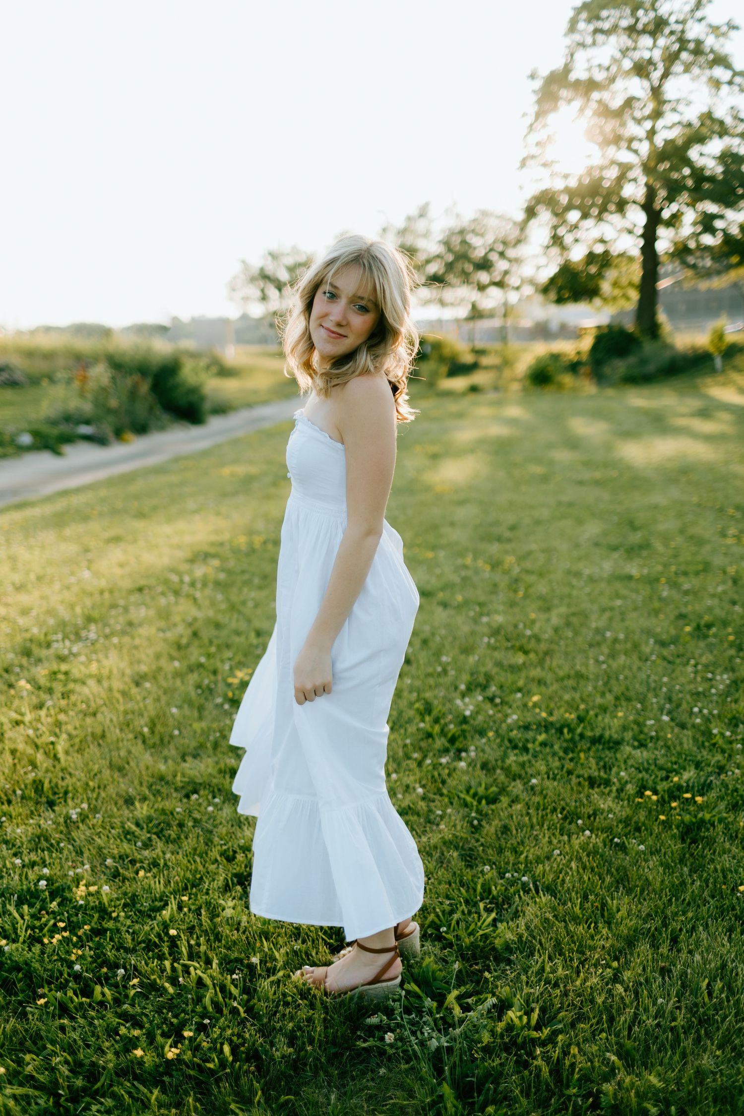 Young blonde woman in white ruffled maxi dress standing on green grass at golden hour sunset.