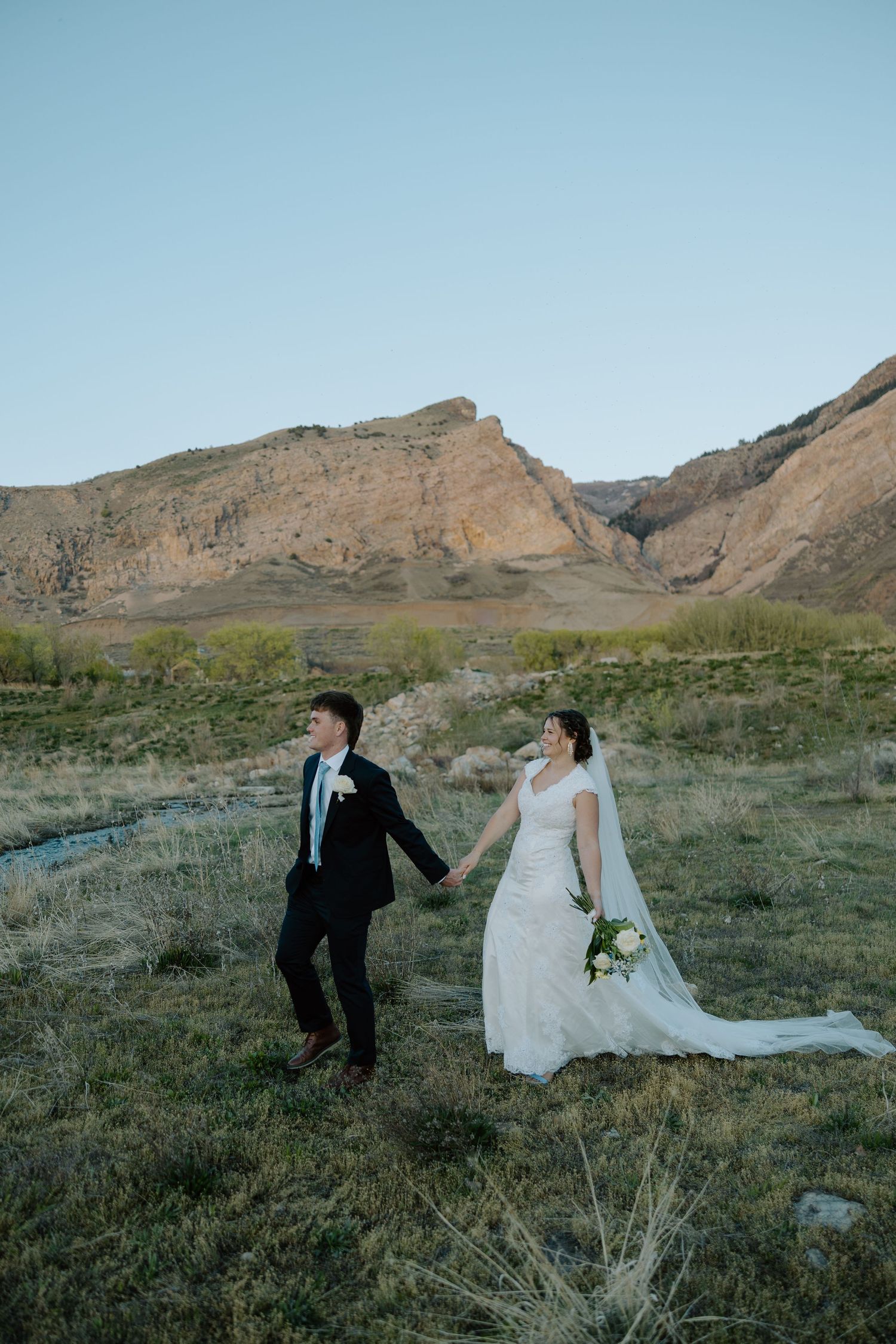 Bride and groom walk hand in hand through a scenic desert field with dramatic mountain landscape.