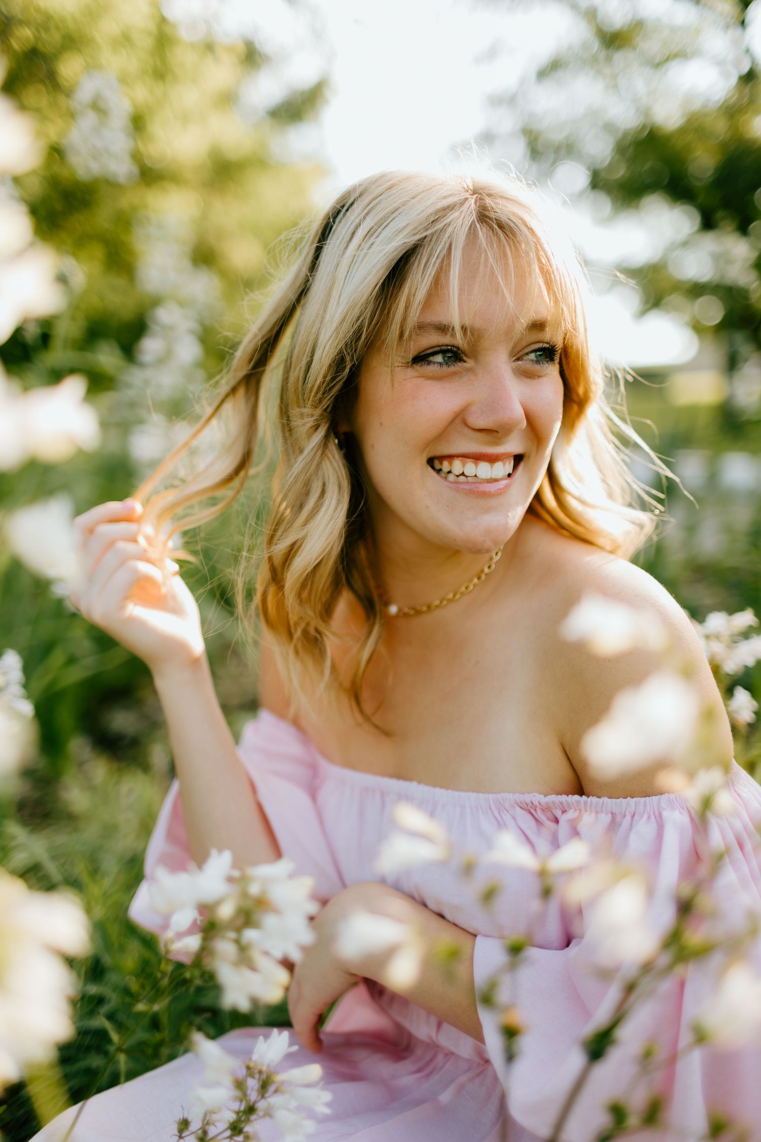 Smiling blonde woman in pink off-shoulder dress sitting among white wildflowers in sunlight.