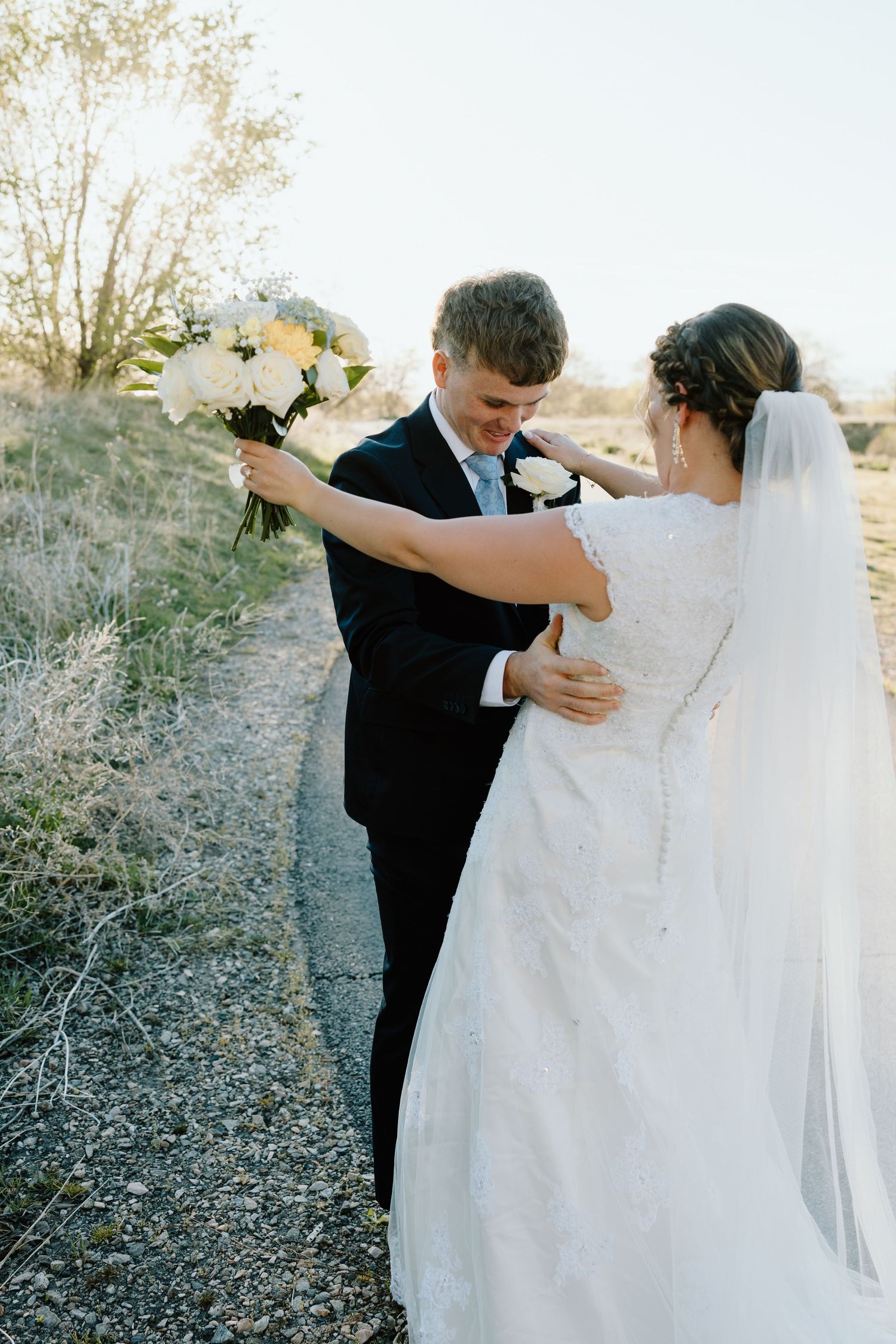 Smiling groom in black suit hands bouquet to bride in lace wedding dress outdoors.