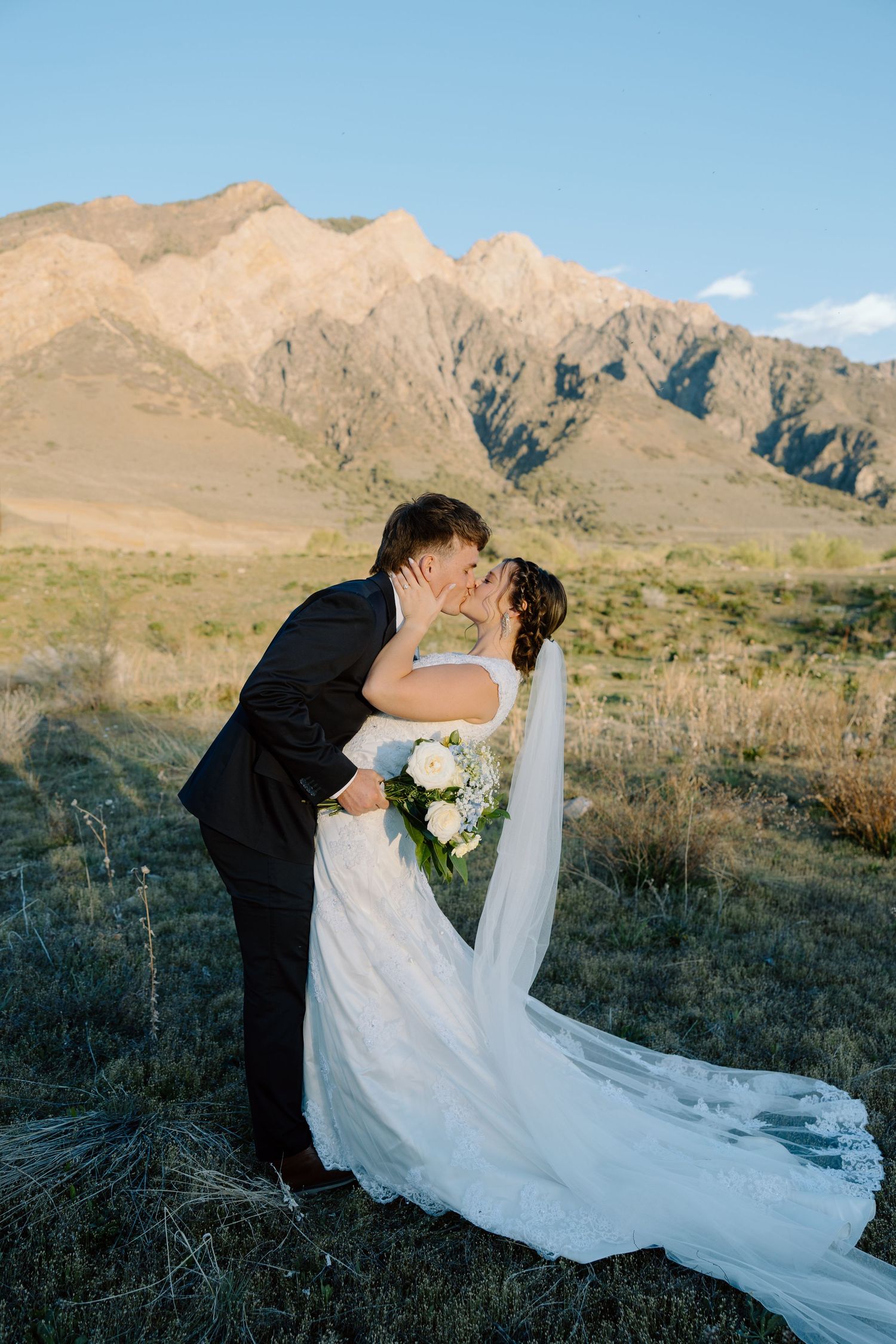 Groom dipping bride for romantic kiss with sunlit rocky mountains in background.