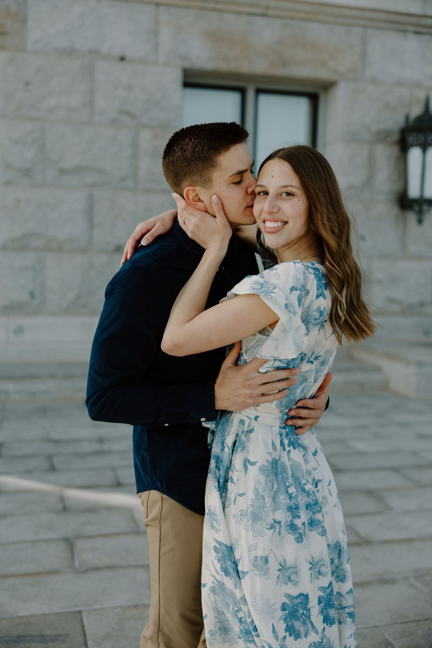 Man in navy shirt kissing smiling woman in blue floral dress on the cheek on marble steps.