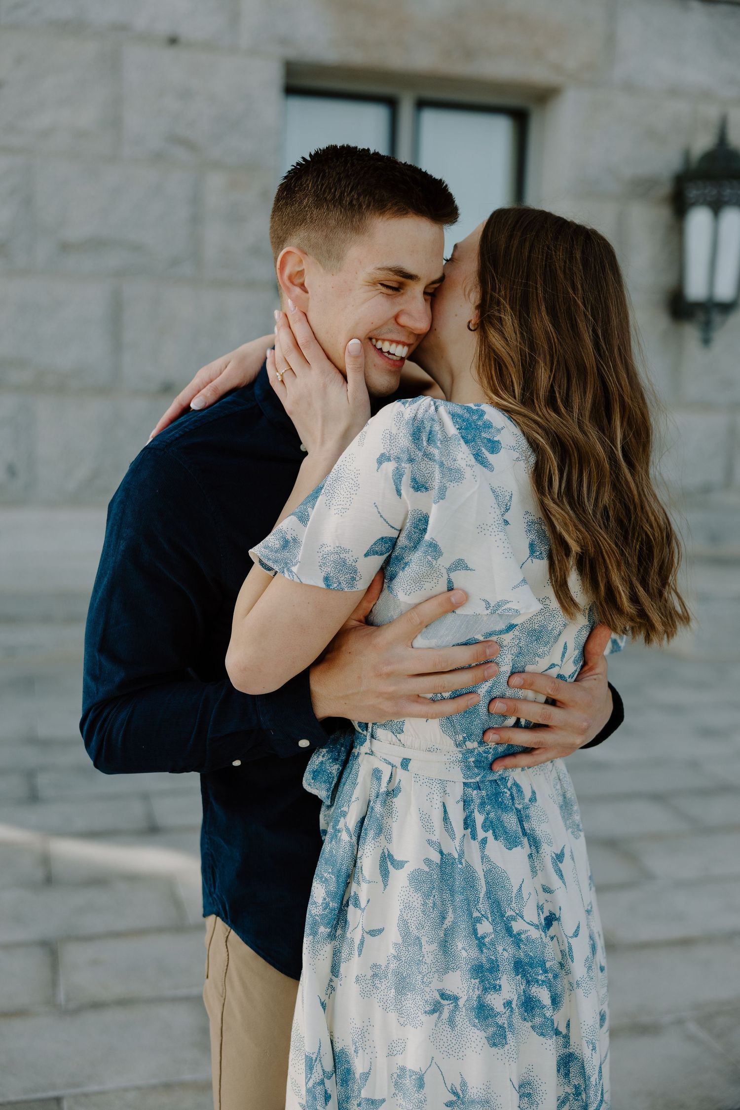 Happy couple sharing a tender moment outside a stone building, woman in blue floral dress embracing man.
