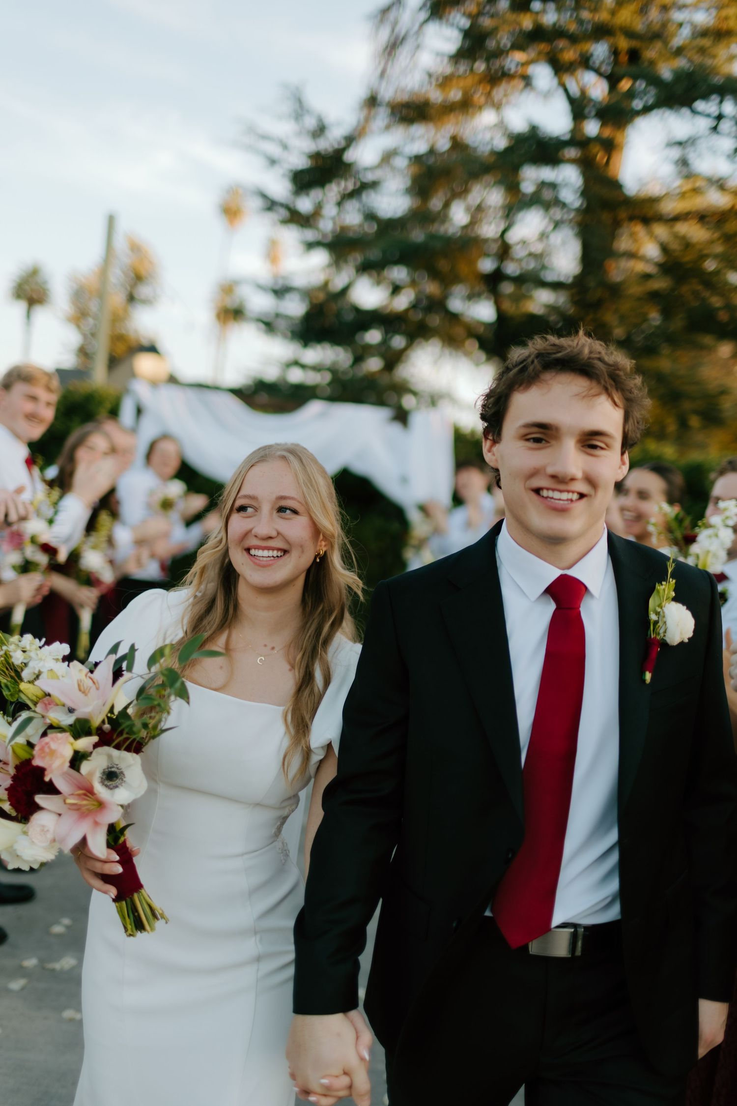 Newlyweds walk hand-in-hand down the aisle, bride holding pink bouquet, groom in black suit with red tie.