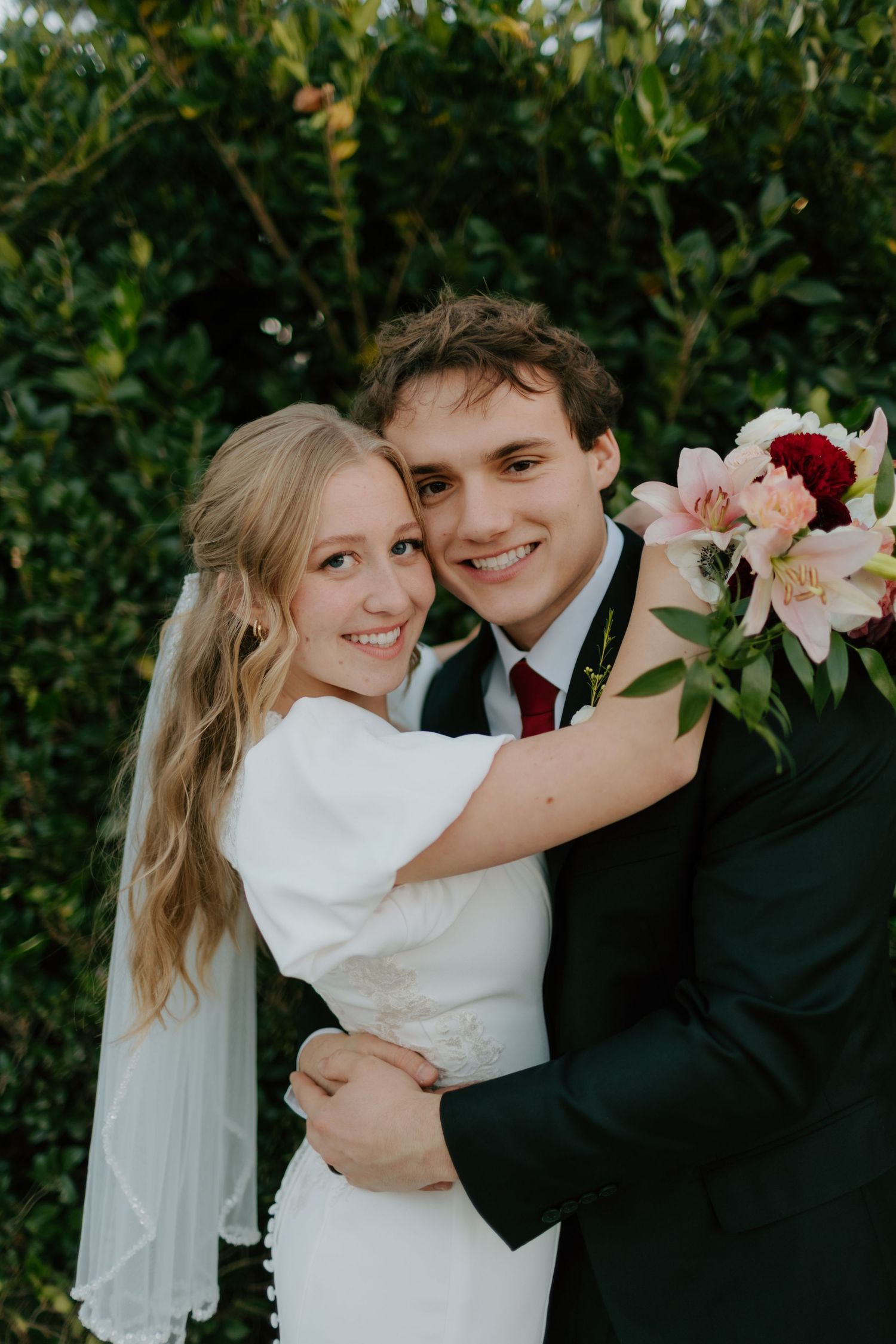 Smiling bride and groom embrace outdoors in front of lush green hedge, bride holding pink and red wedding bouquet.