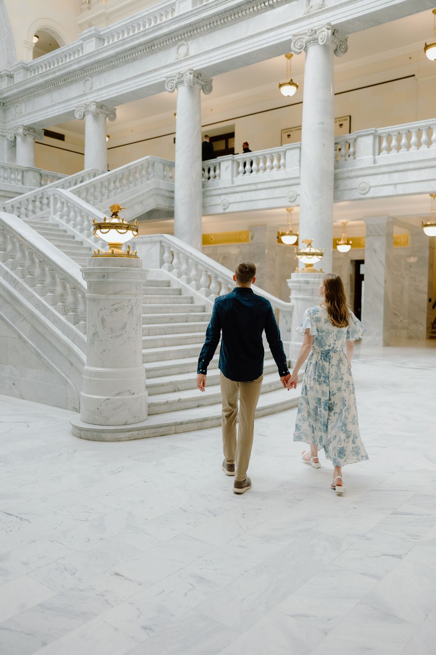 Couple holding hands walking toward grand marble staircase in elegant white columned building interior.