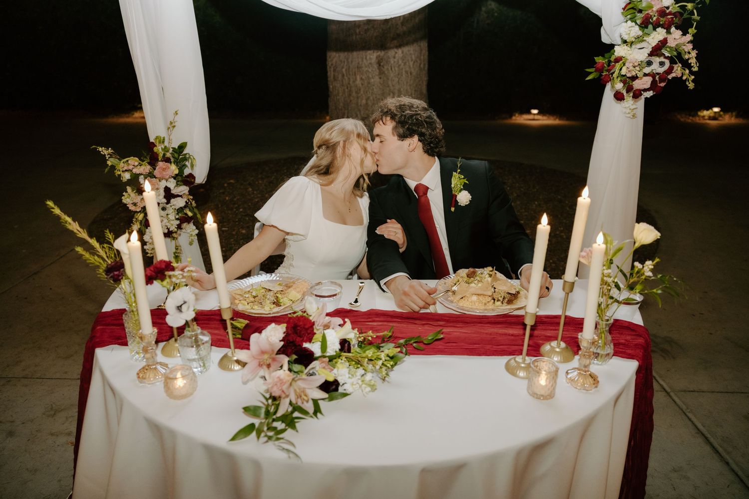 Bride and groom share a kiss at candlelit sweetheart table with burgundy runner and floral centerpieces.