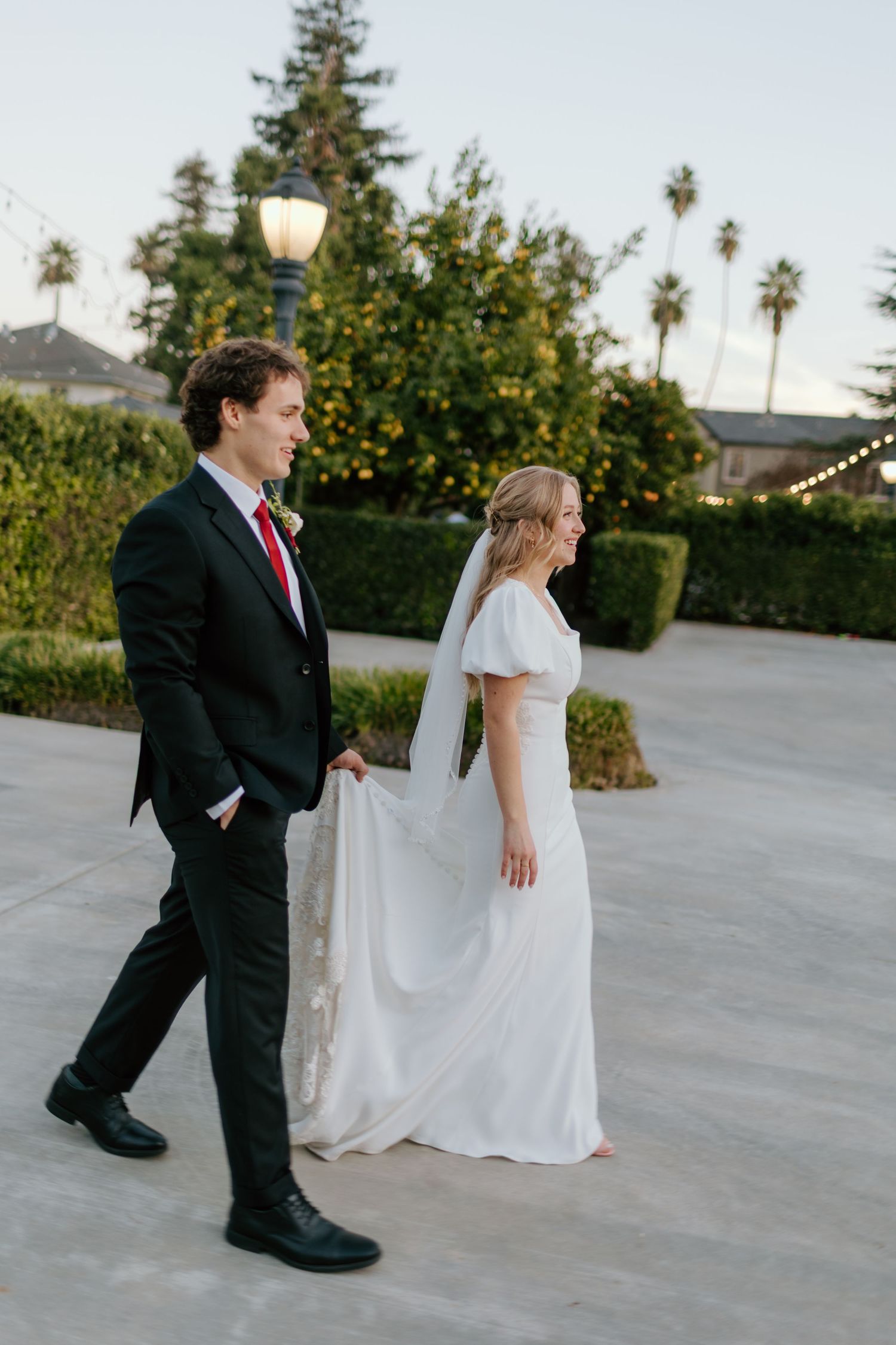 Bride in white dress with veil and groom in dark suit with red tie walking together on outdoor patio with palm trees backdrop.