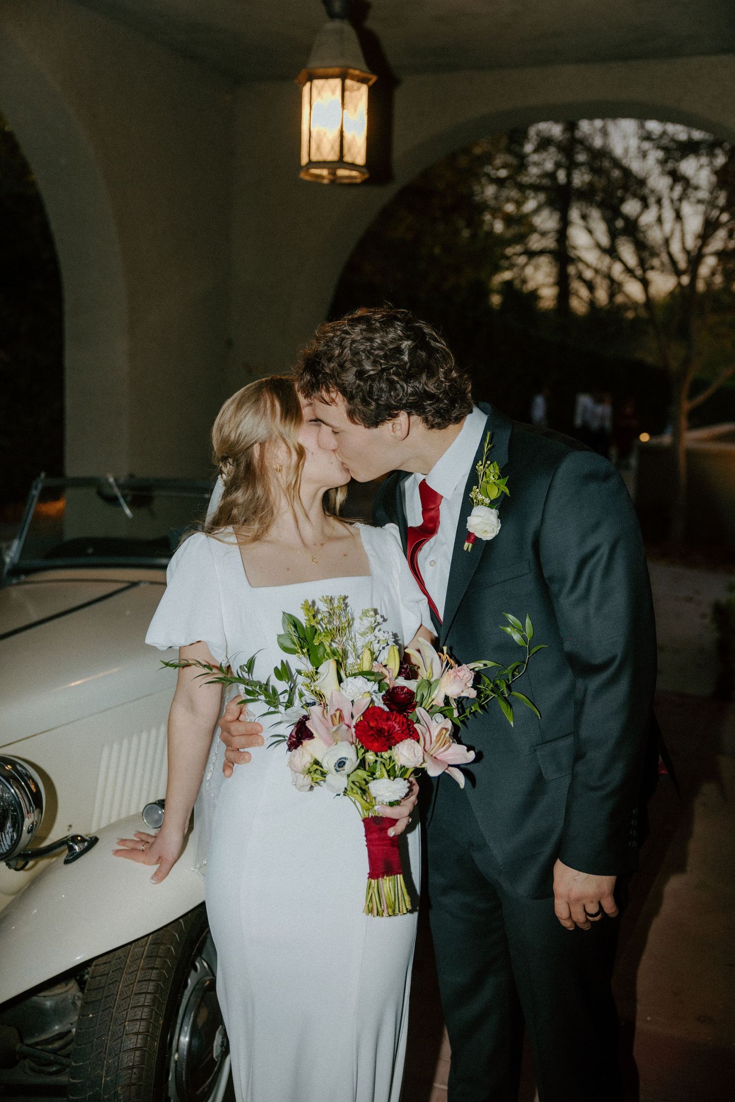 Bride and groom sharing a kiss beside a vintage white car, holding a red and pink floral bouquet at dusk.