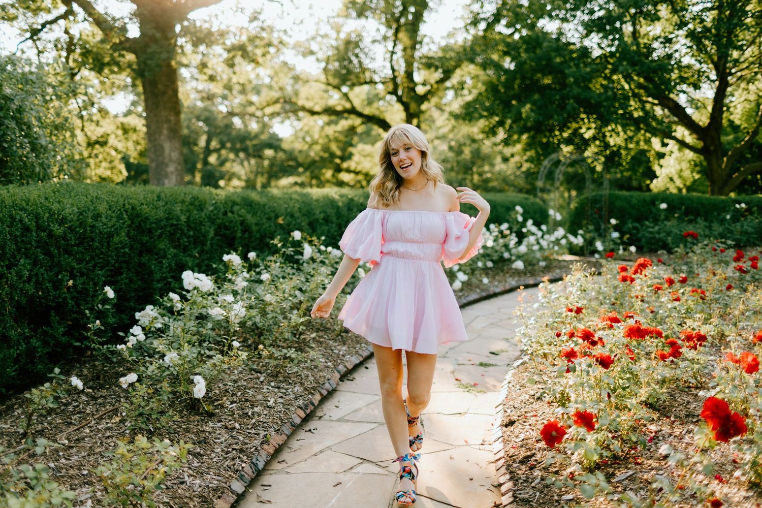 Woman in pink off-shoulder dress walks garden path among red and white roses with lush green trees.