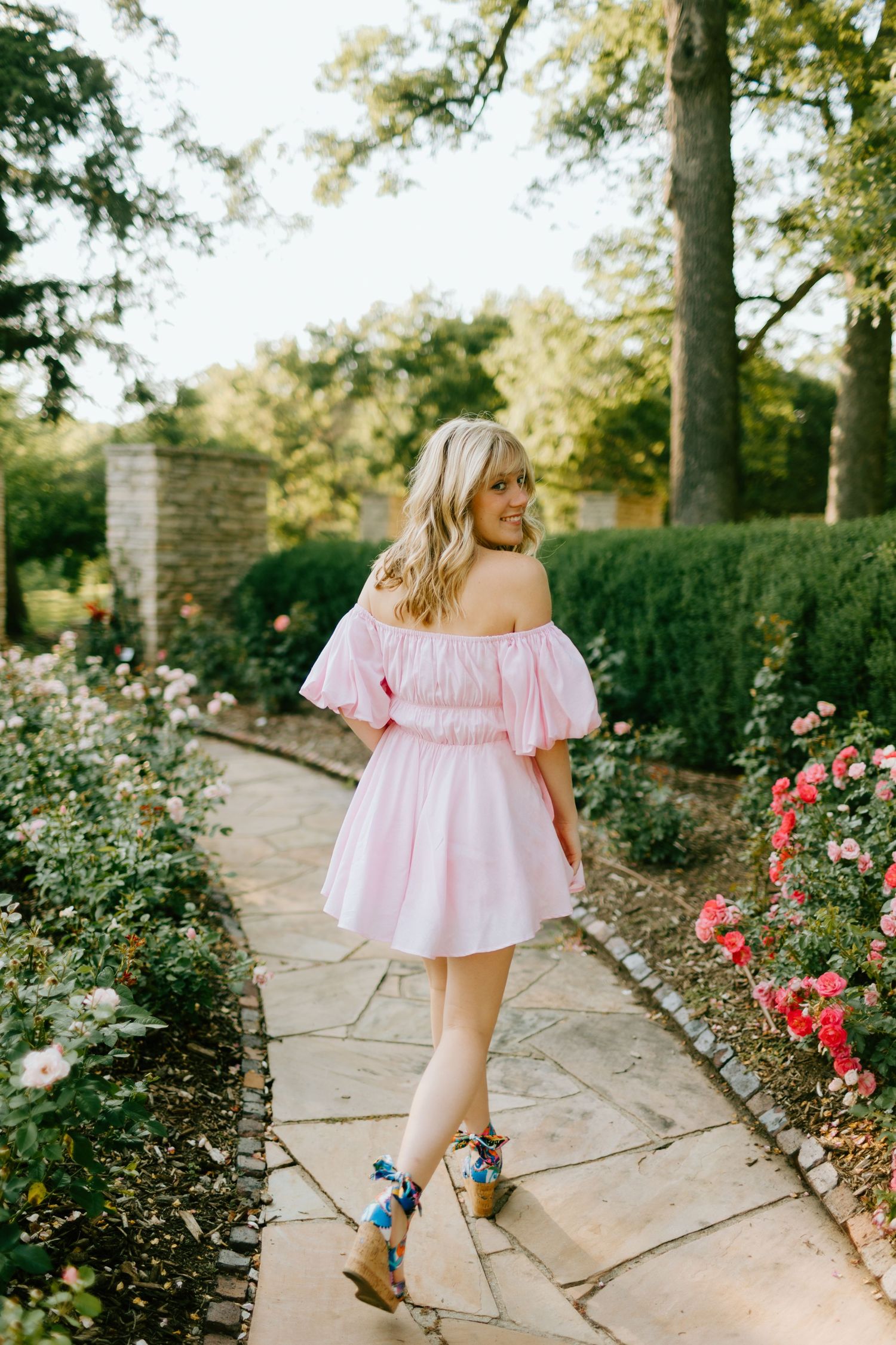 Woman in pink puff-sleeve off-shoulder dress strolling through rose garden on stone pathway.