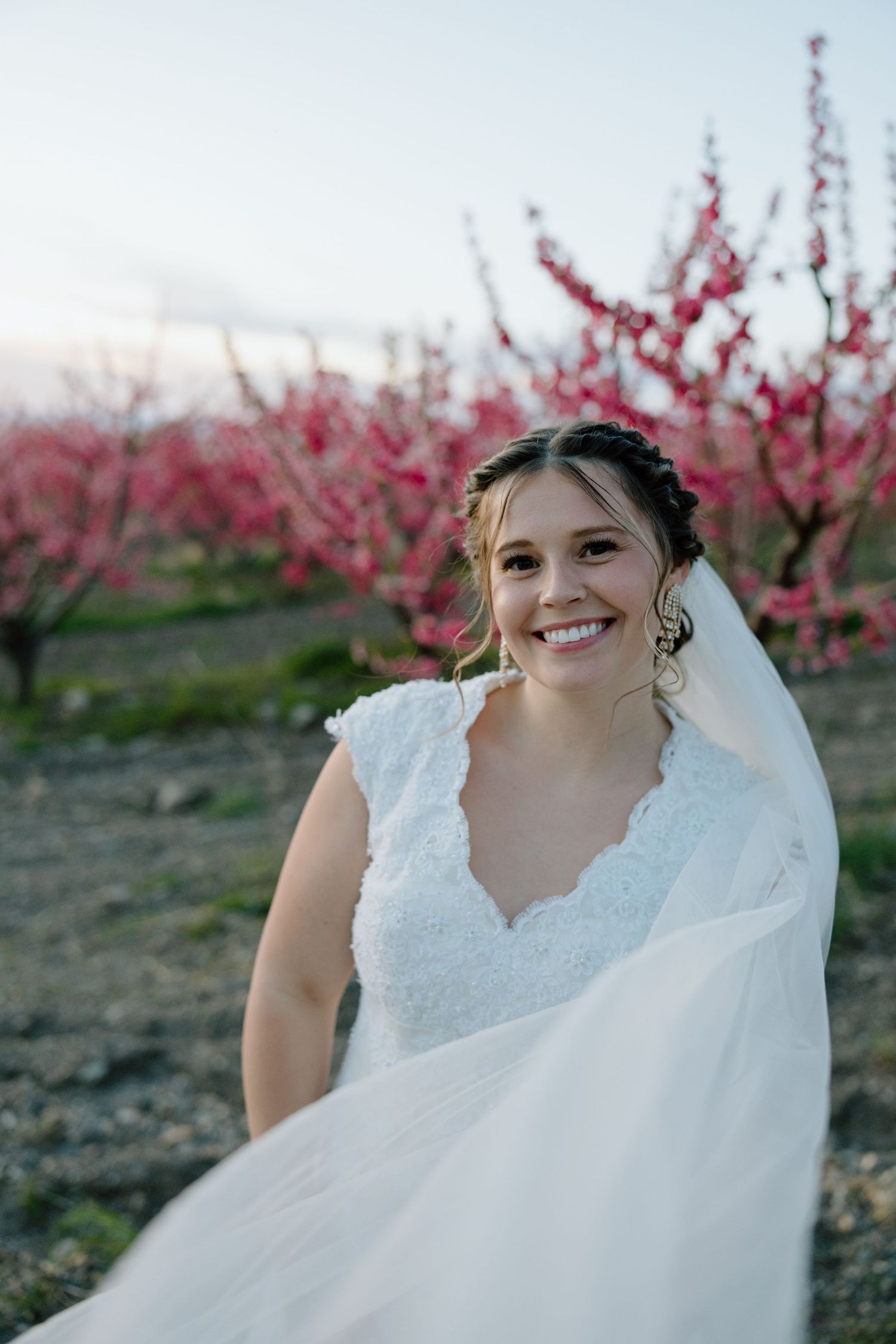 Radiant bride smiles beautifully in a lace wedding dress with pink flowering trees in background.
